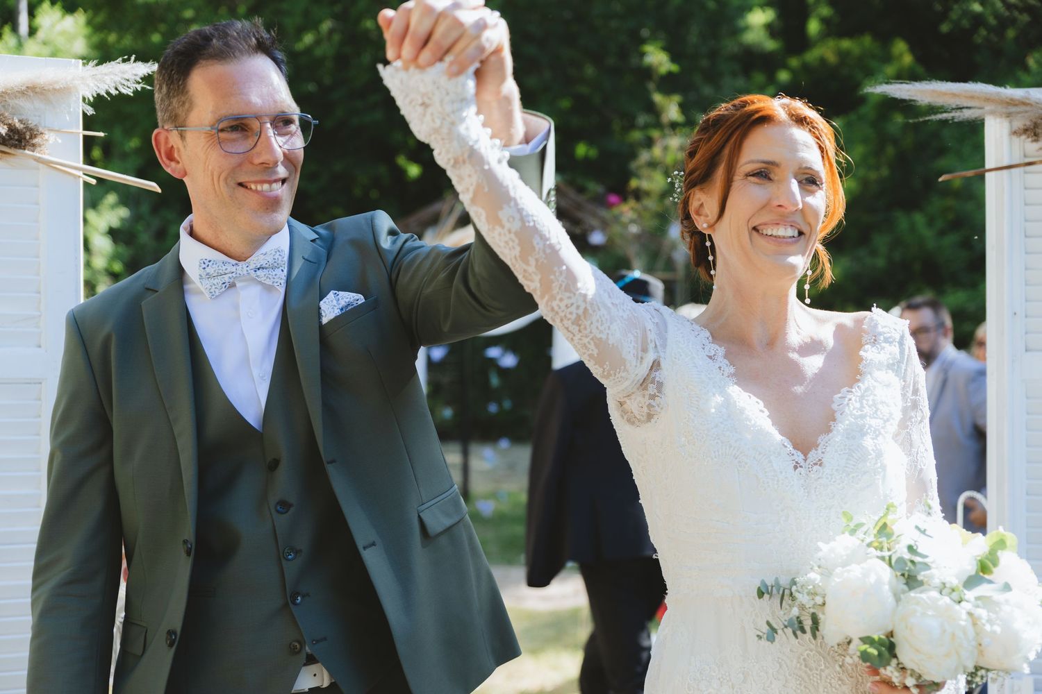 A joyful newlywed couple celebrating their wedding ceremony, groom in green suit holding bride's hand up in triumph.