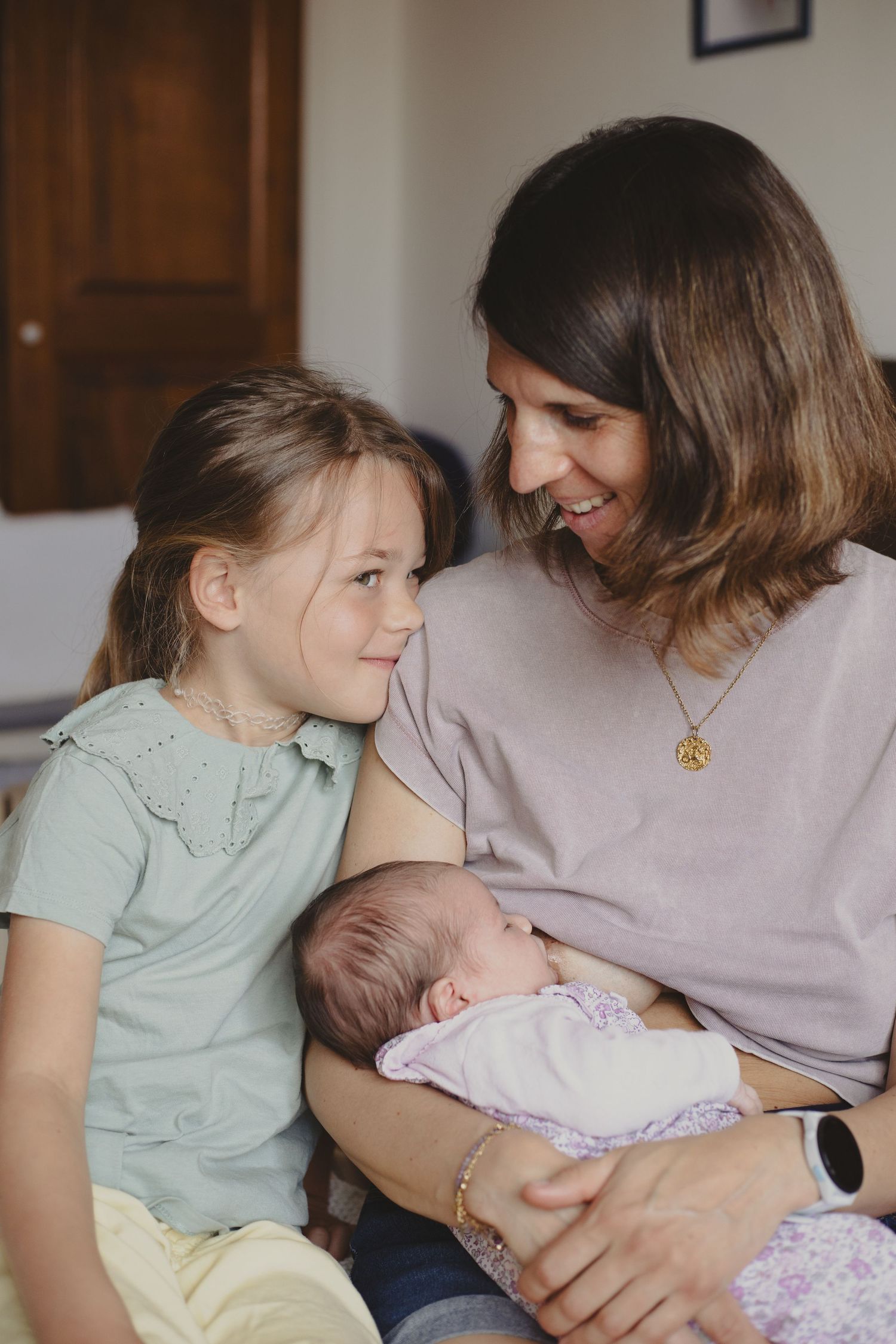 Smiling mother with brown hair holding a newborn while her young daughter looks up at her.