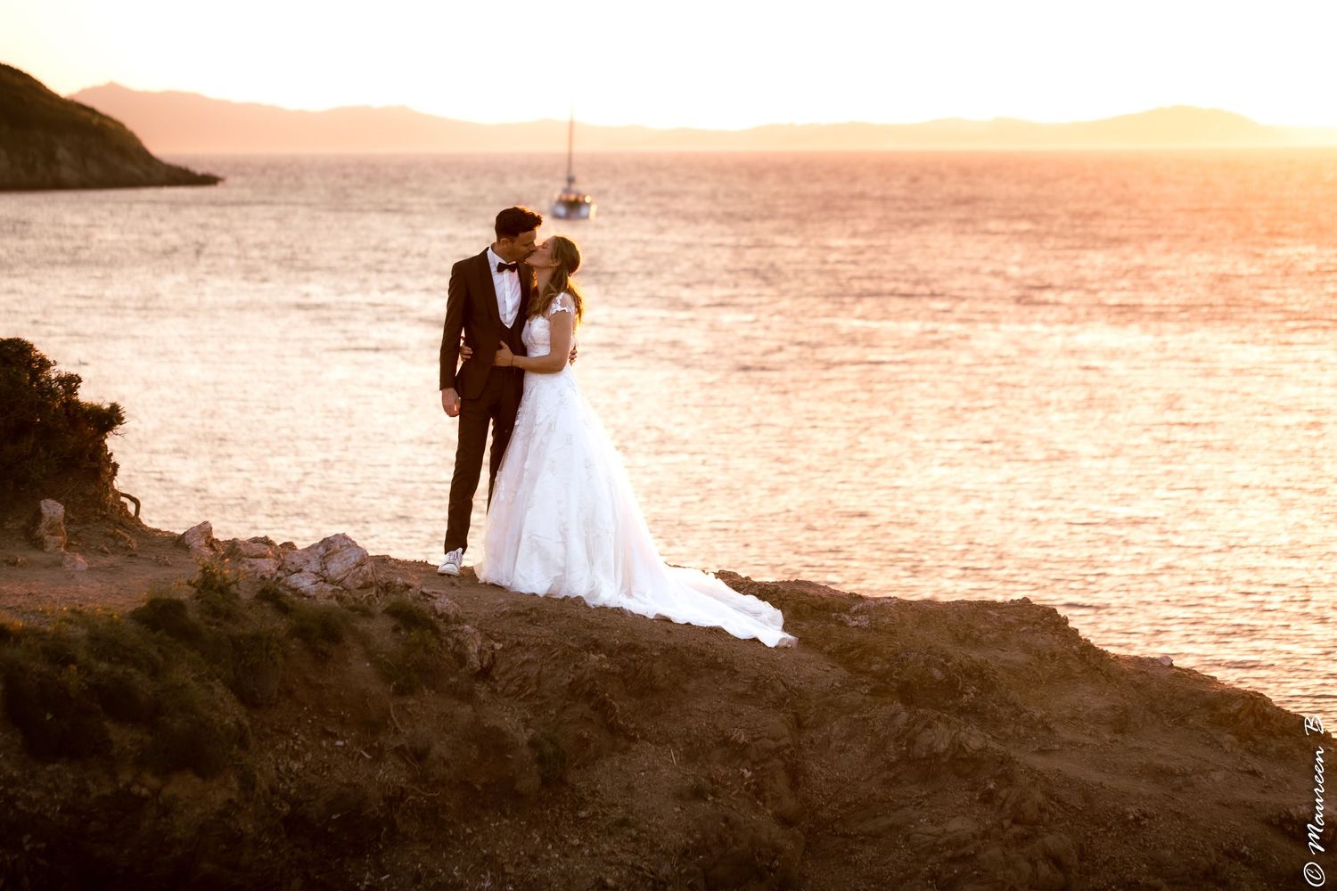 Bride and groom kissing on rocky coastline at sunset with sailboat on golden water behind them.