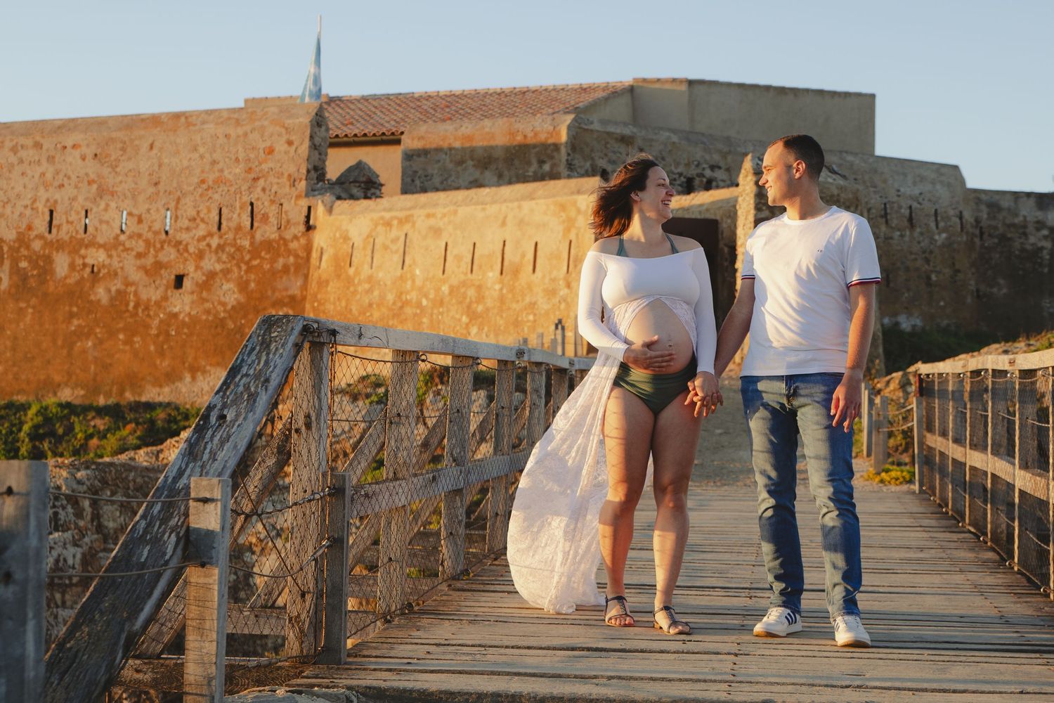 Pregnant woman and partner walking on wooden bridge near historic fortress at sunset, woman in white lace overlay.