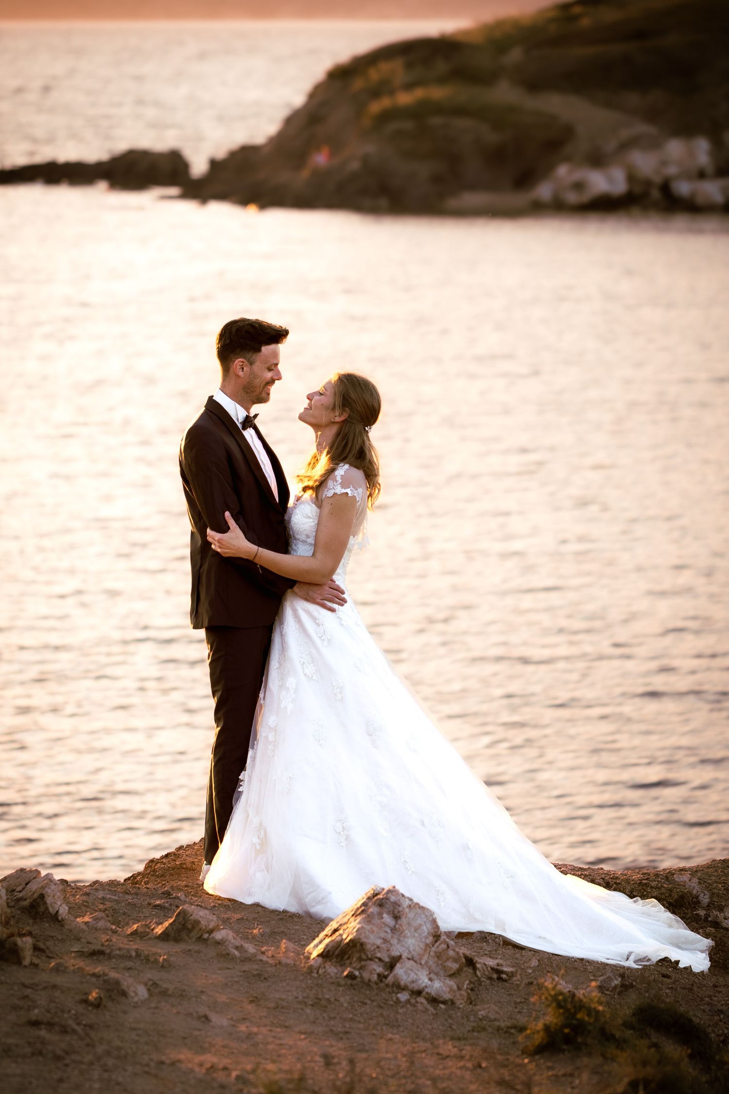 Bride and groom embrace romantically at sunset by the ocean, with golden light reflecting on the water.
