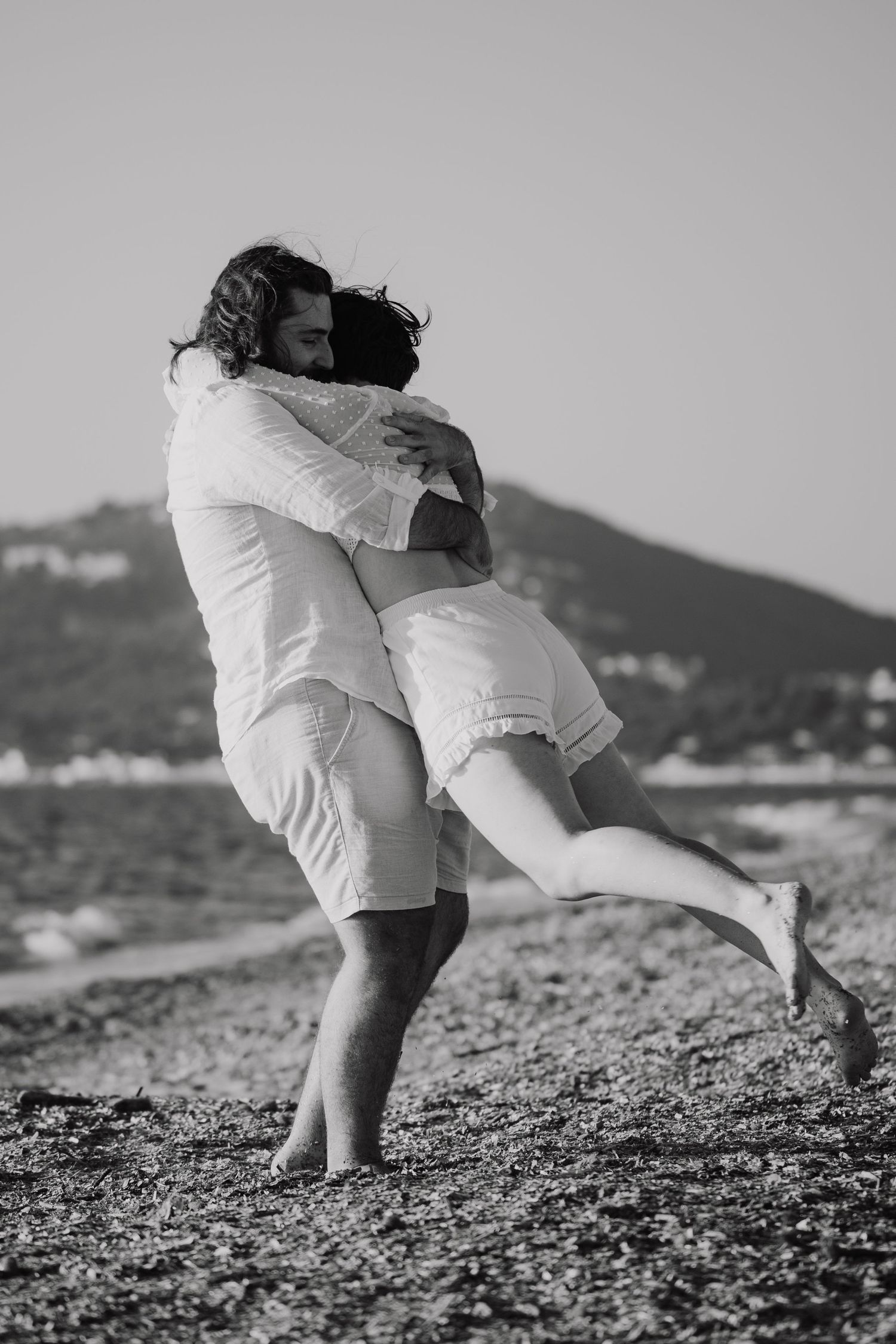Black and white photo of a couple embracing on a rocky beach, man lifting woman in a romantic hug.