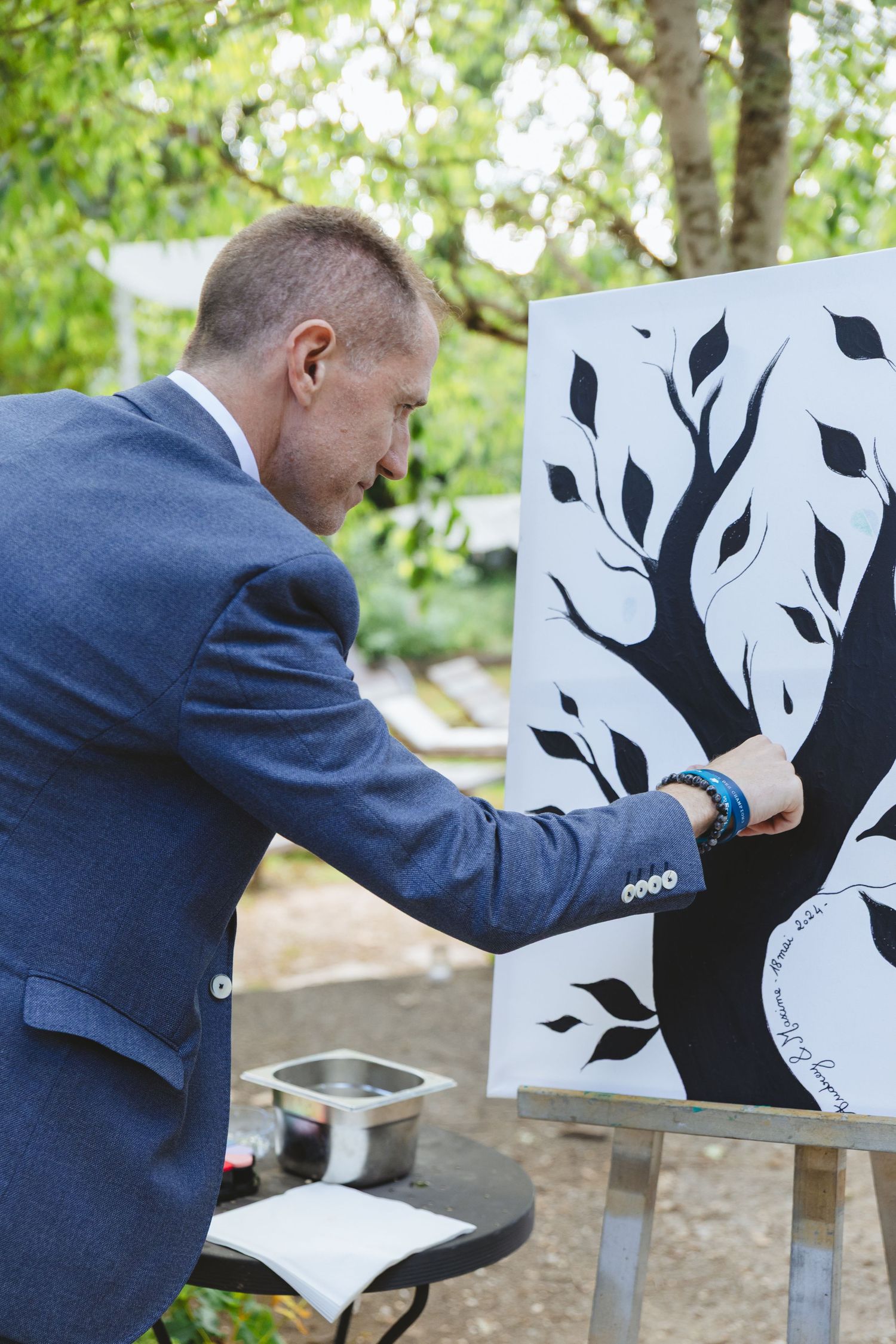 Man in blue suit signing a black and white tree canvas on an easel at an outdoor wedding reception.