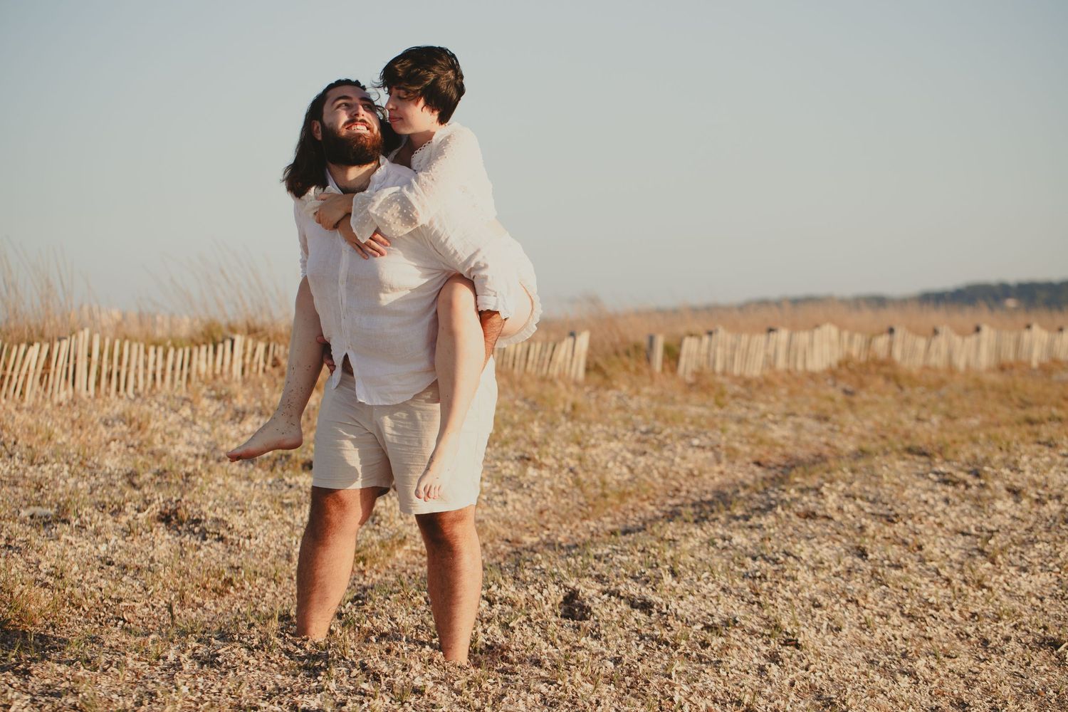 A couple enjoys a piggyback ride on a sandy beach, smiling and dressed in casual neutral outfits on a sunny day.