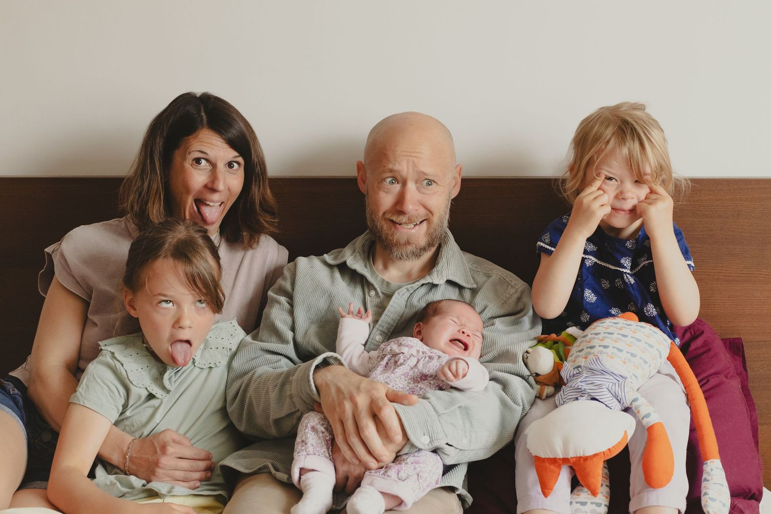 Playful family photo with parents making funny faces while holding their newborn and children.