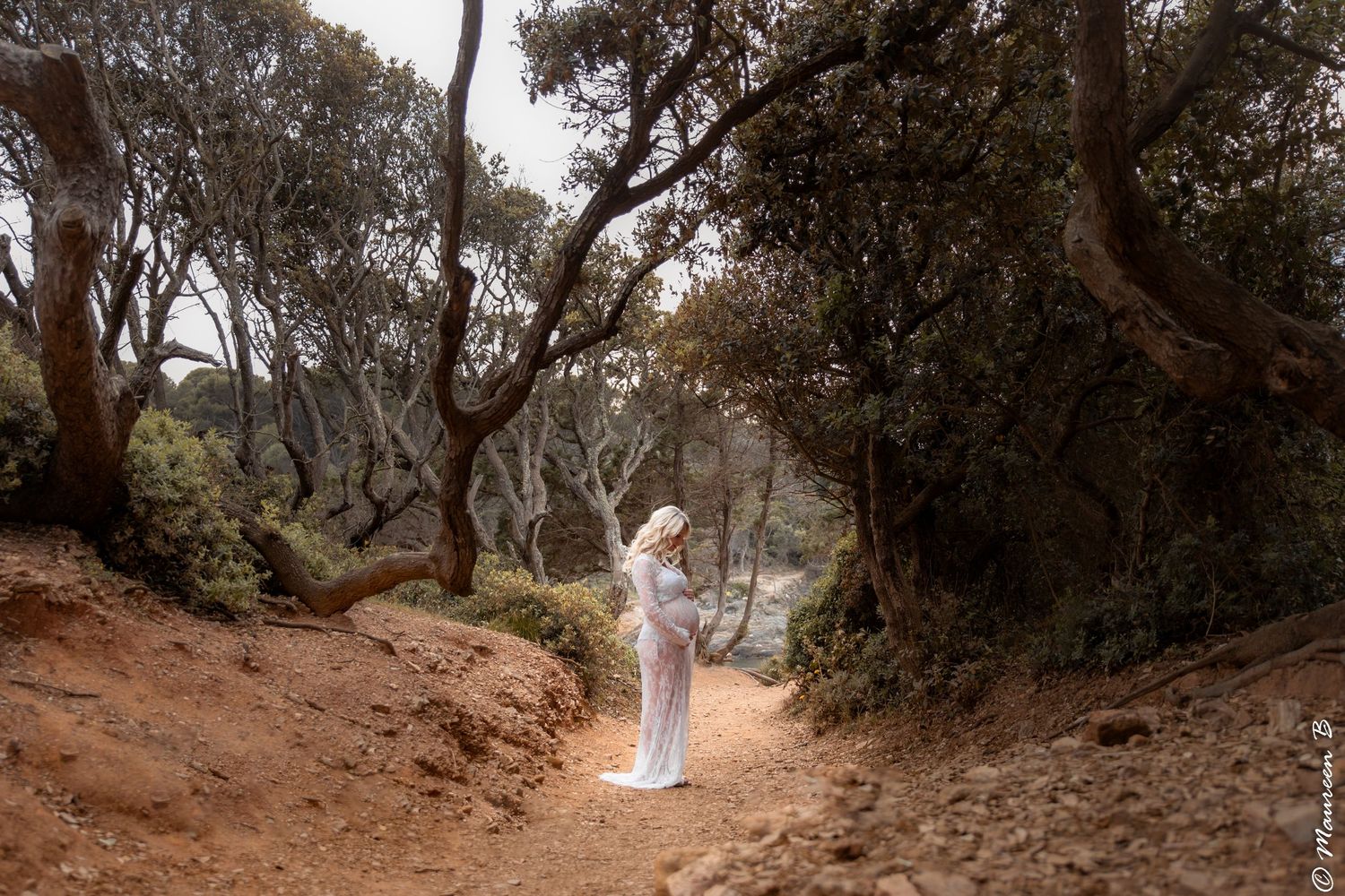 Pregnant woman in white dress standing on a scenic forest path among twisted trees.