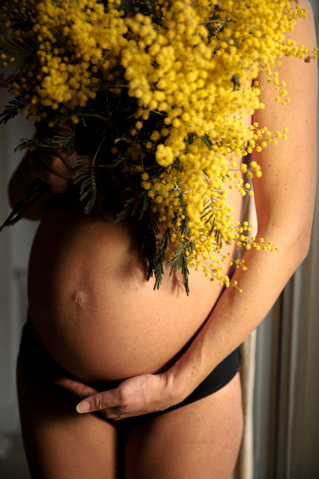 Pregnant woman holding yellow mimosa flowers over her bare belly in warm light.