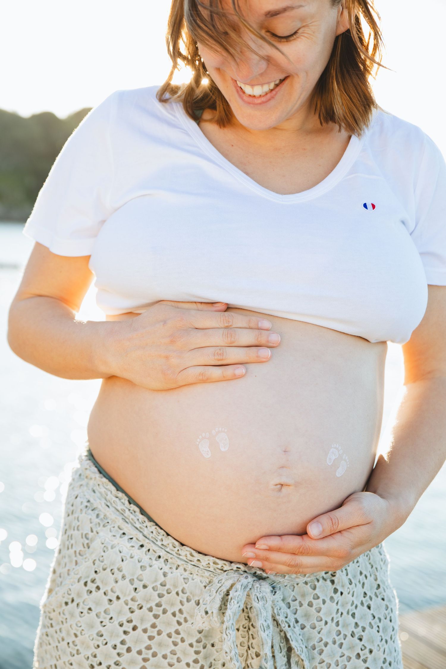 Smiling pregnant woman in white tee applying cream to bare belly outdoors near water in warm sunlight.