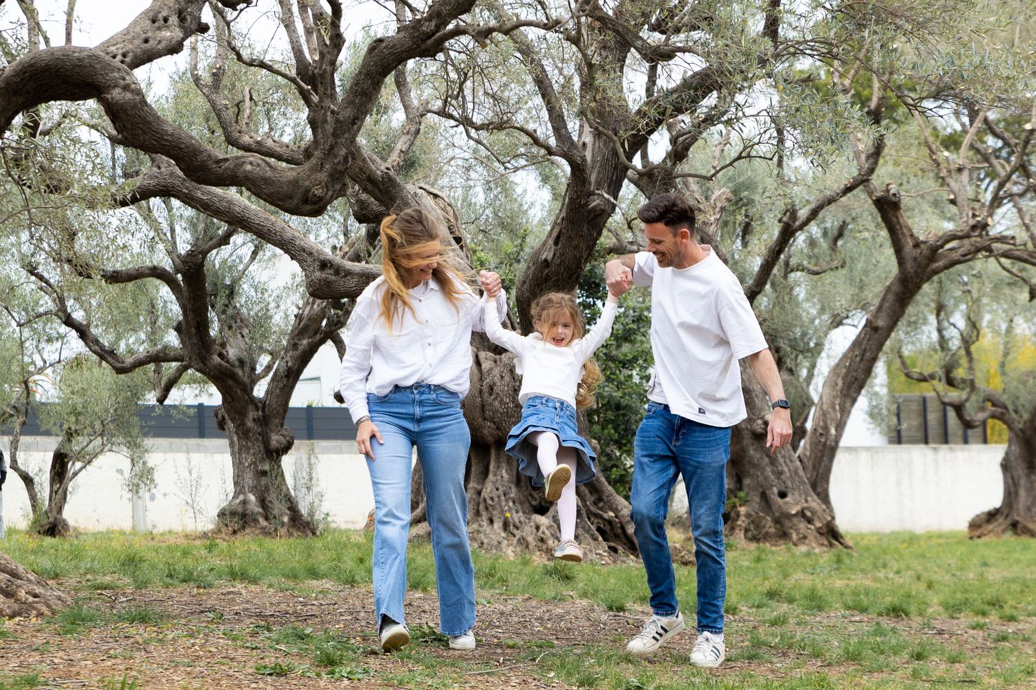 A happy family swings their young daughter between them while walking outdoors among ancient olive trees.