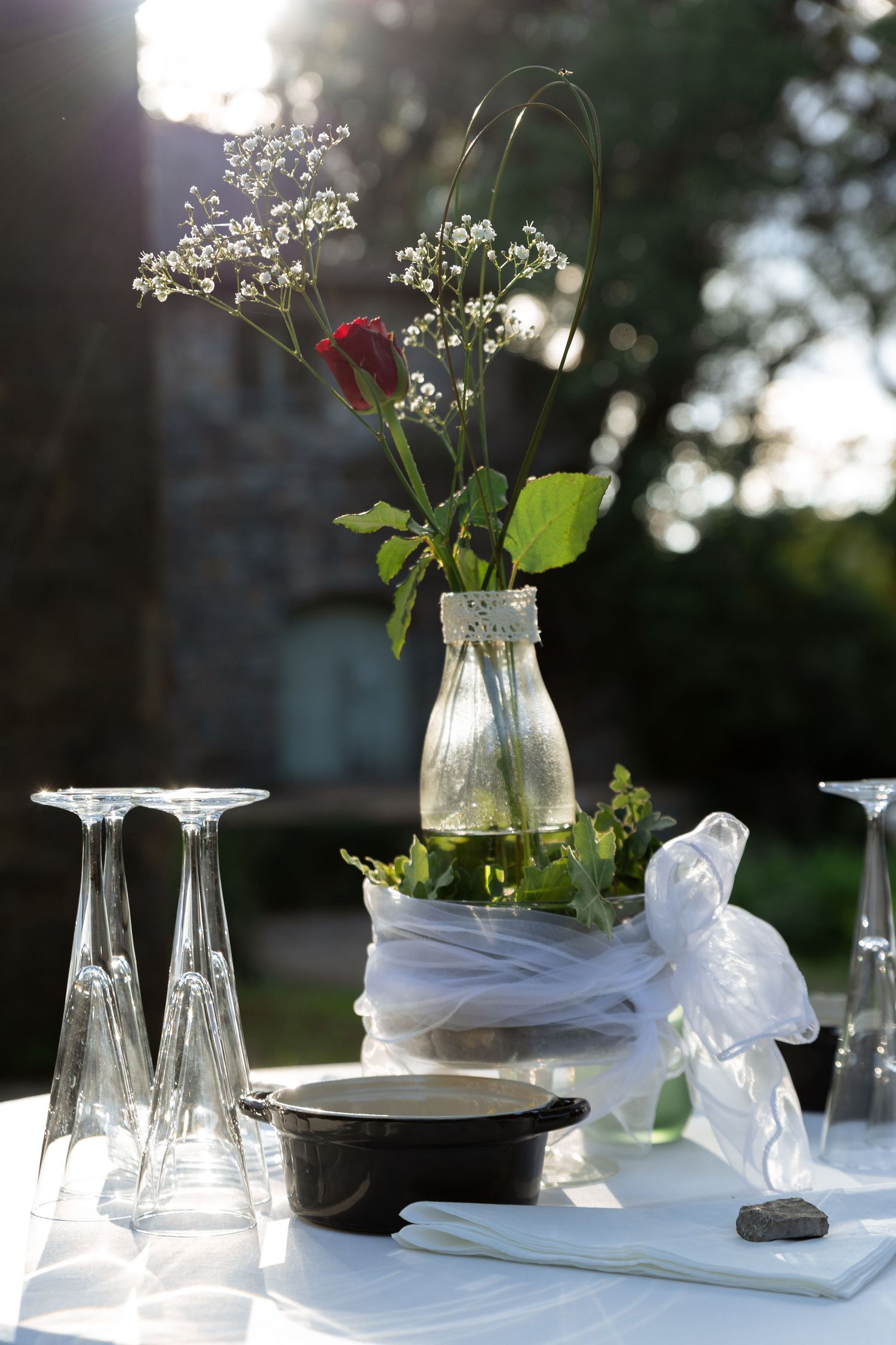 Outdoor table setting with glass vases holding red roses and baby's breath, wine glasses, and a small bowl on white linen.
