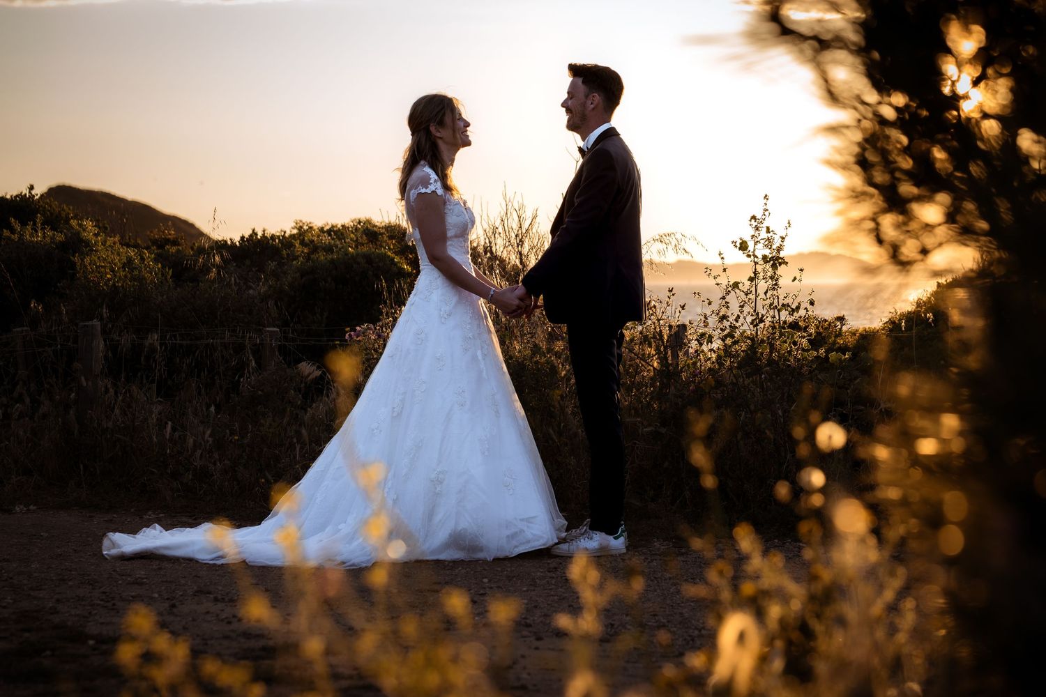 Bride and groom hold hands at golden sunset outdoors, bride in white gown, groom in dark suit.