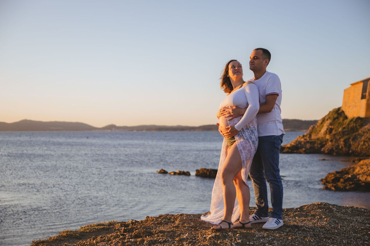 Expecting couple standing together on rocky shore at golden hour, embracing with scenic sea and hills behind them.