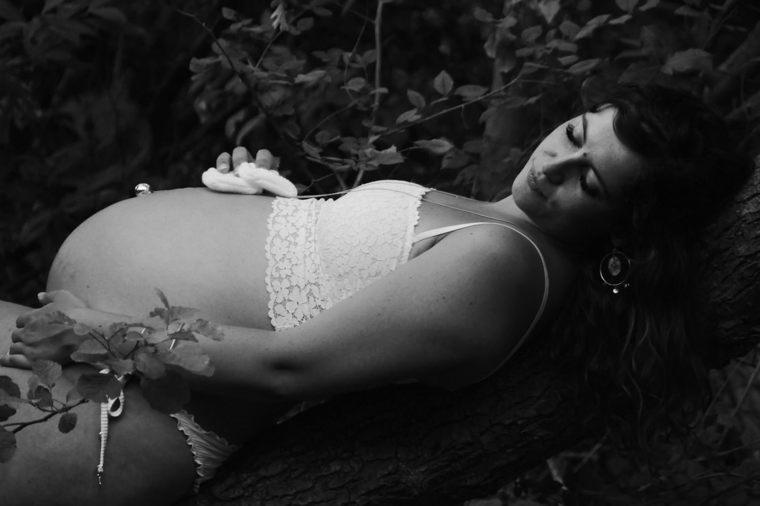 Black and white photo of pregnant woman in white lace lingerie resting among leaves outdoors.