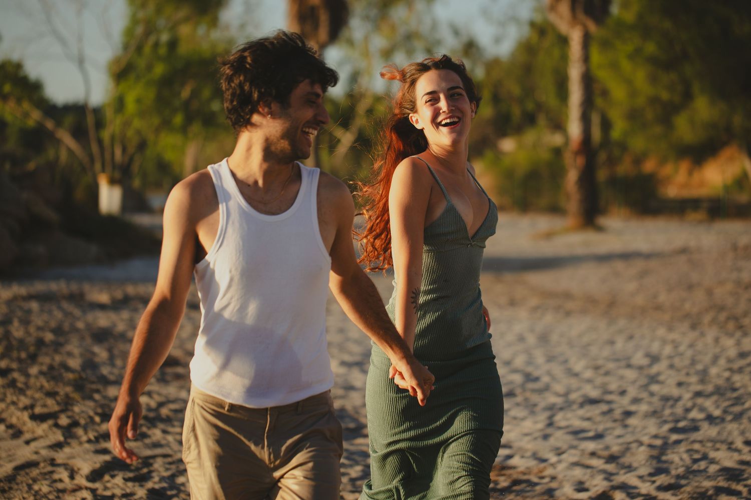 A happy young couple holds hands and laughs while walking on a sandy beach at golden hour.
