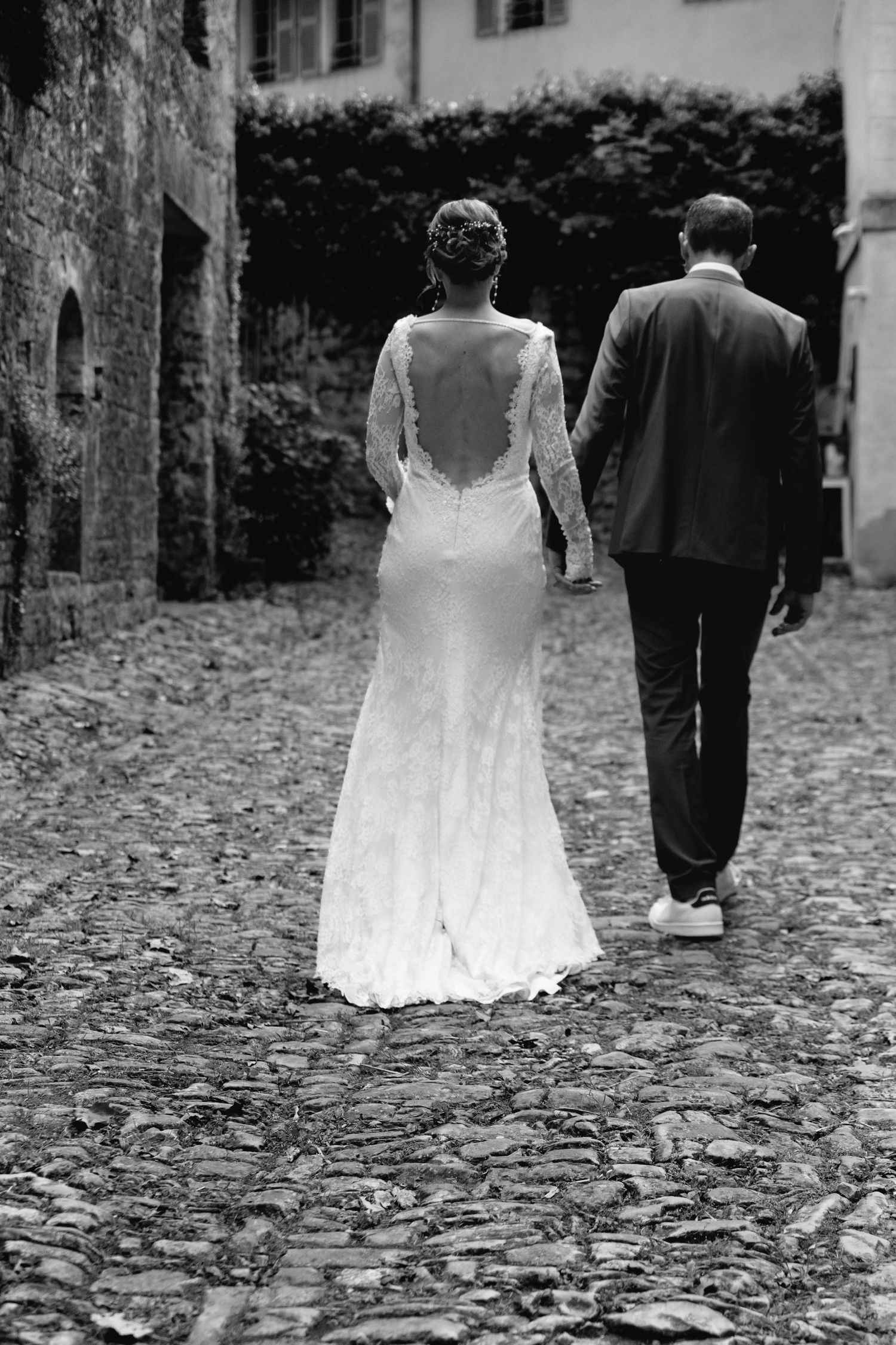 Black and white photo of bride and groom walking hand in hand on cobblestone street in elegant wedding attire.