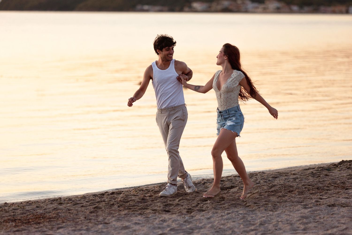 Happy young couple holding hands and running along a sandy beach at golden sunset.
