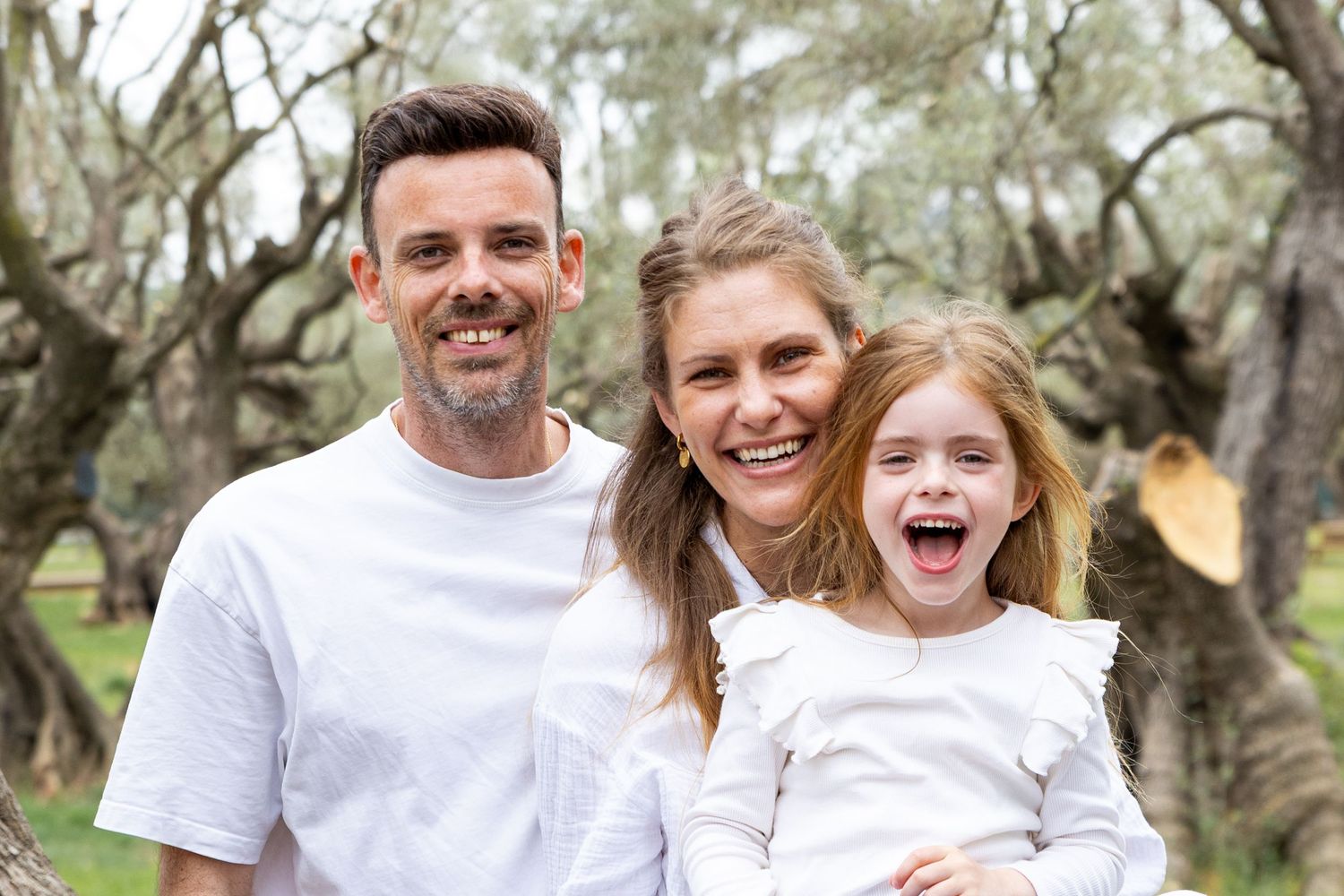 A smiling family of three poses together outdoors among olive trees, with their daughter laughing joyfully.