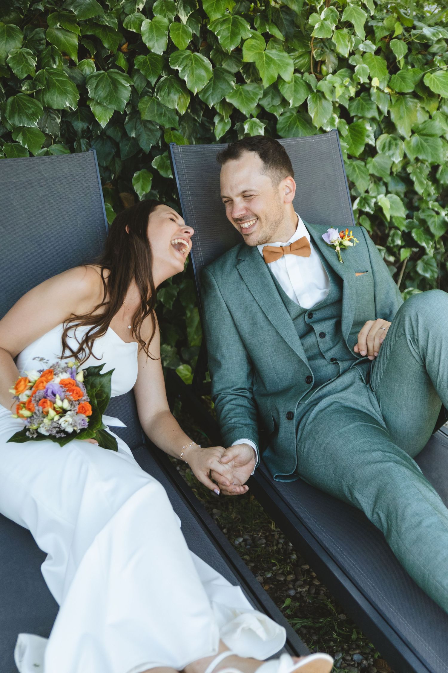 Bride and groom laugh together holding hands while relaxing on lounge chairs against an ivy-covered wall.