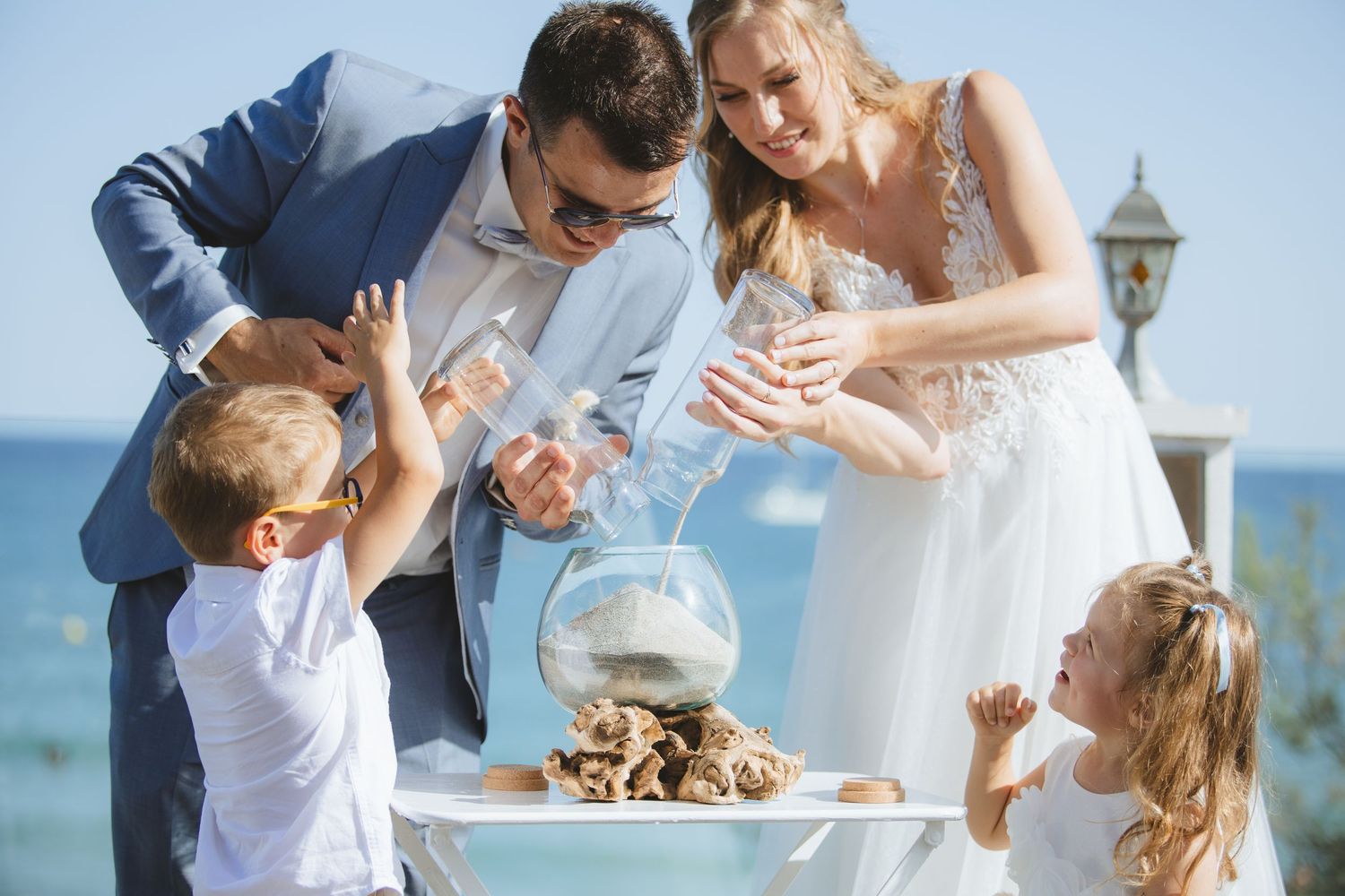 Bride and groom performing sand ceremony with two young children by the sea on wedding day.