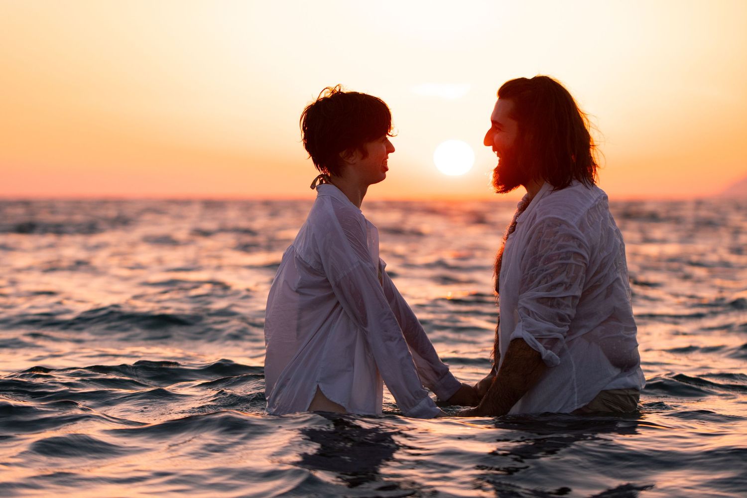 Two people holding hands in the ocean at sunset, wearing white shirts, sharing a romantic moment.
