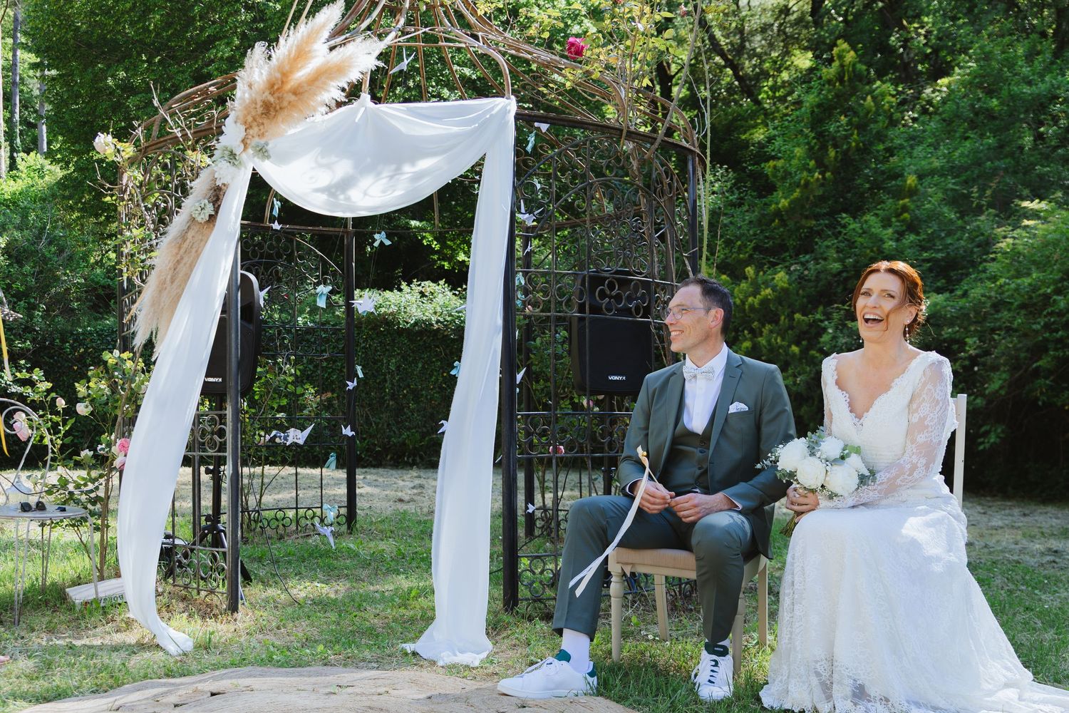 Bride and groom laughing together outdoors at a garden wedding ceremony with a white draped floral arch.