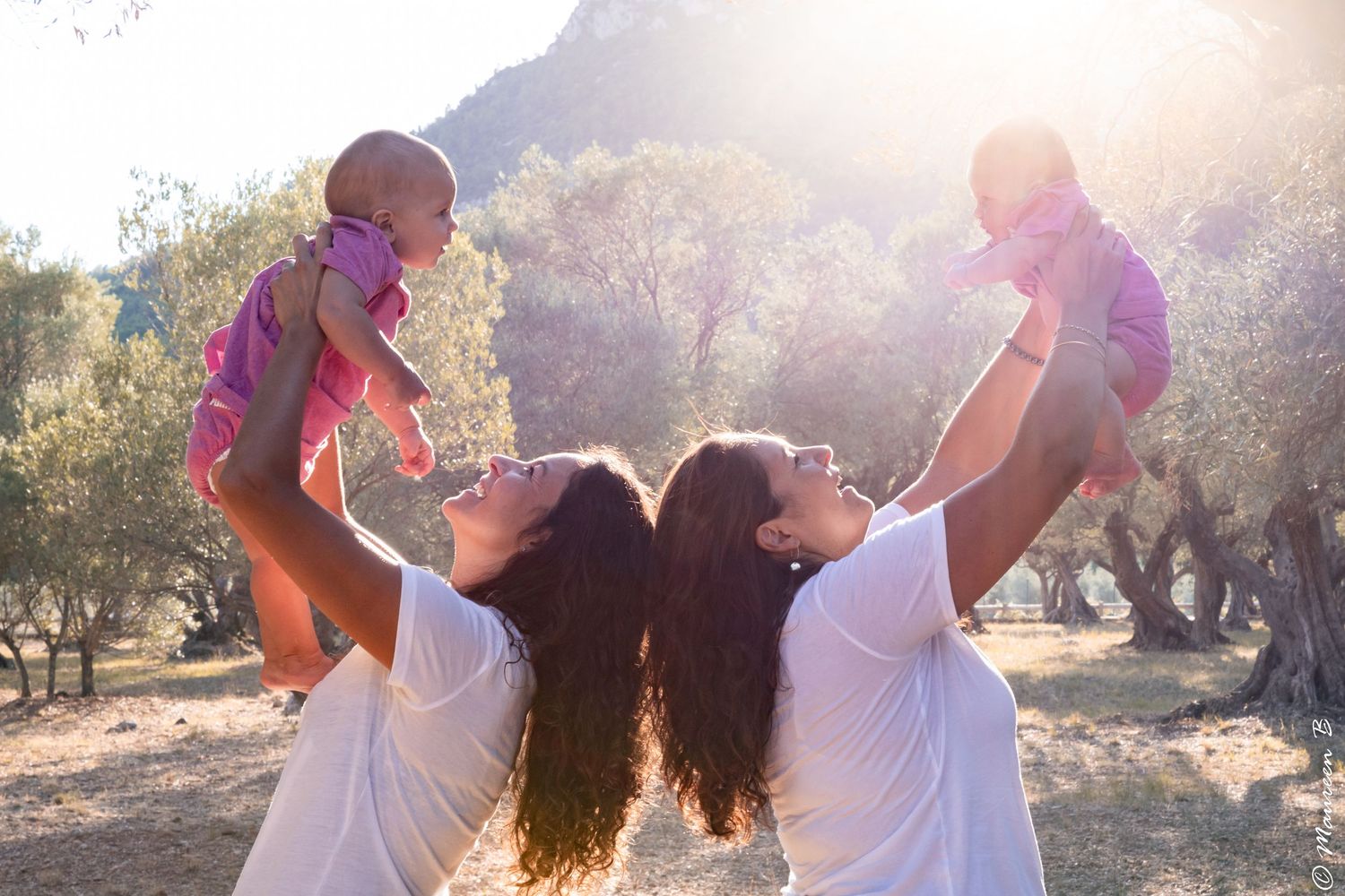 Two mothers in white shirts joyfully lift their babies in a sunlit olive grove.