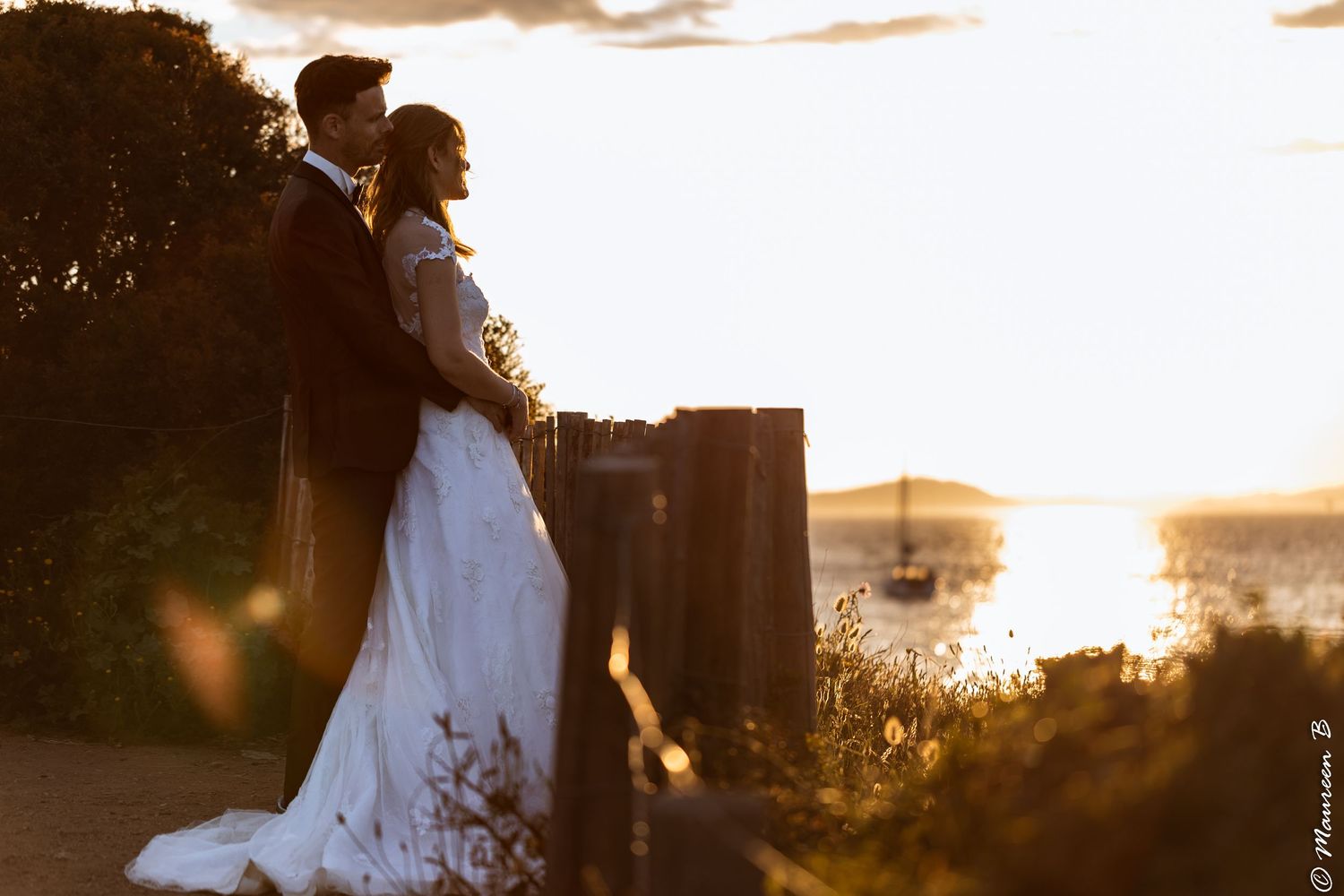 Bride and groom embracing at waterfront fence with sailboat and stunning golden sunset behind them.