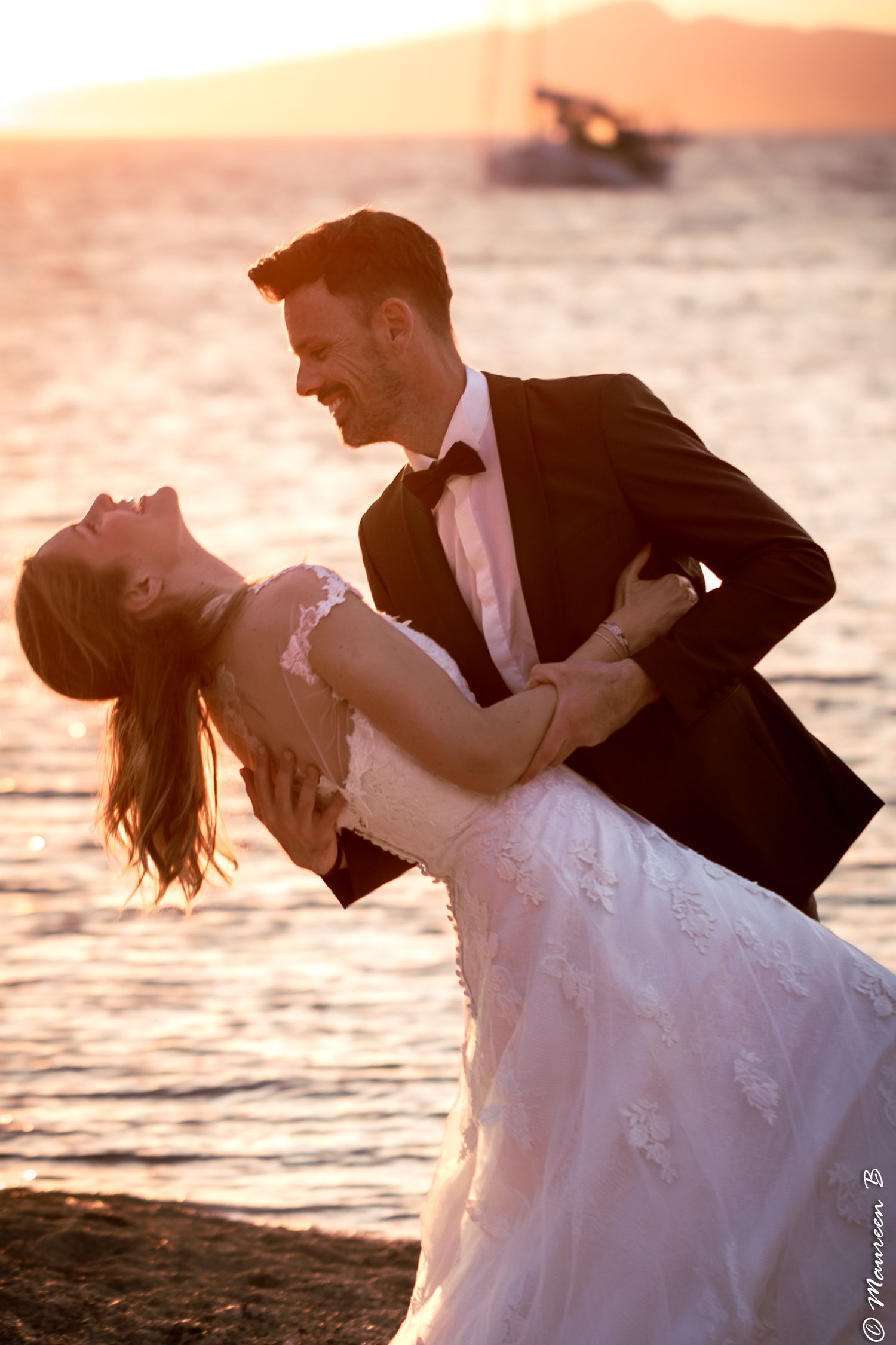Groom dipping bride dramatically on beach at sunset, both laughing joyfully together.