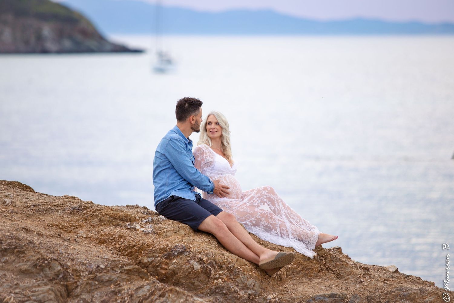 Loving couple seated on coastal rocks, pregnant woman in white lace dress smiling during maternity shoot.