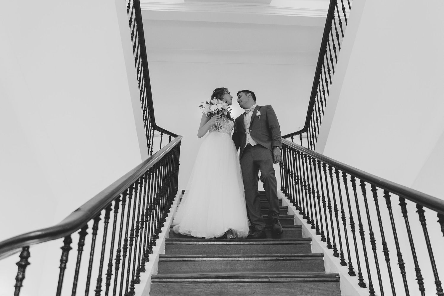 Black and white photo of newlywed couple kissing on elegant staircase with ornate railings.