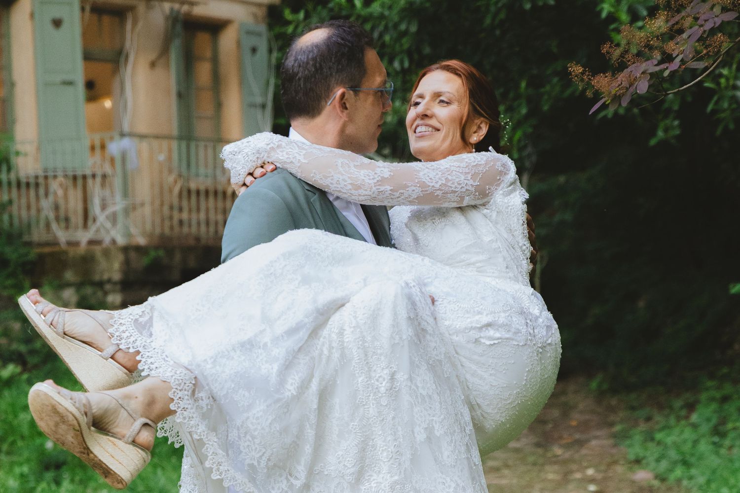 Groom carrying bride in white lace dress outdoors, both smiling joyfully on their wedding day.