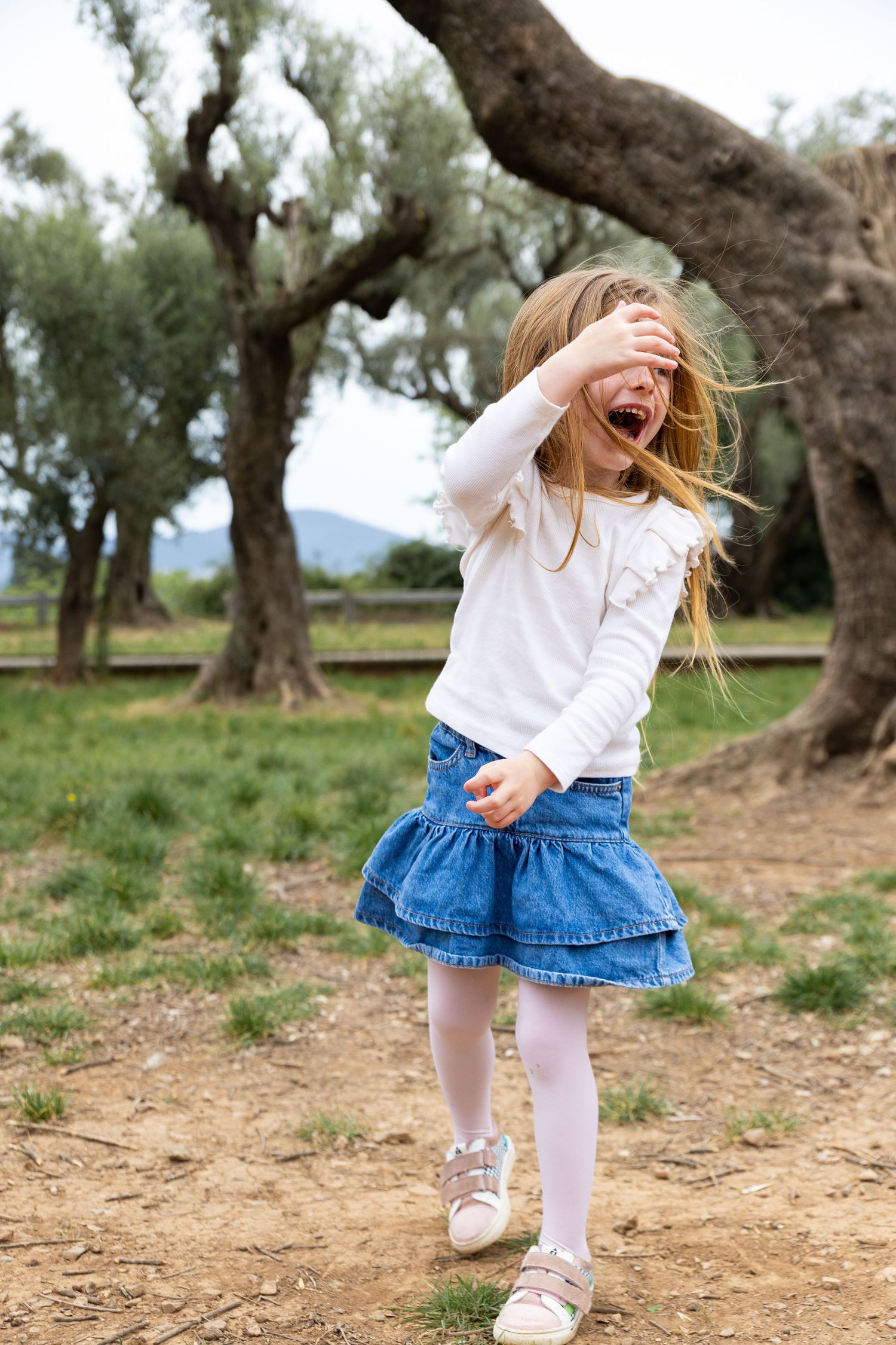 A young blonde girl in a white top and denim skirt laughs and covers her eye outdoors.