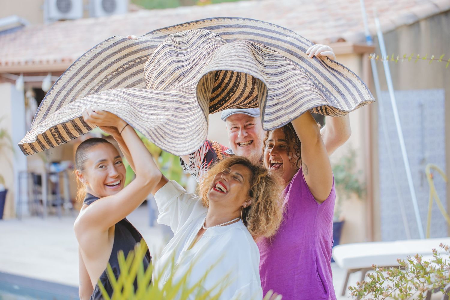A cheerful group of people huddle under a large striped hat outdoors near a pool.