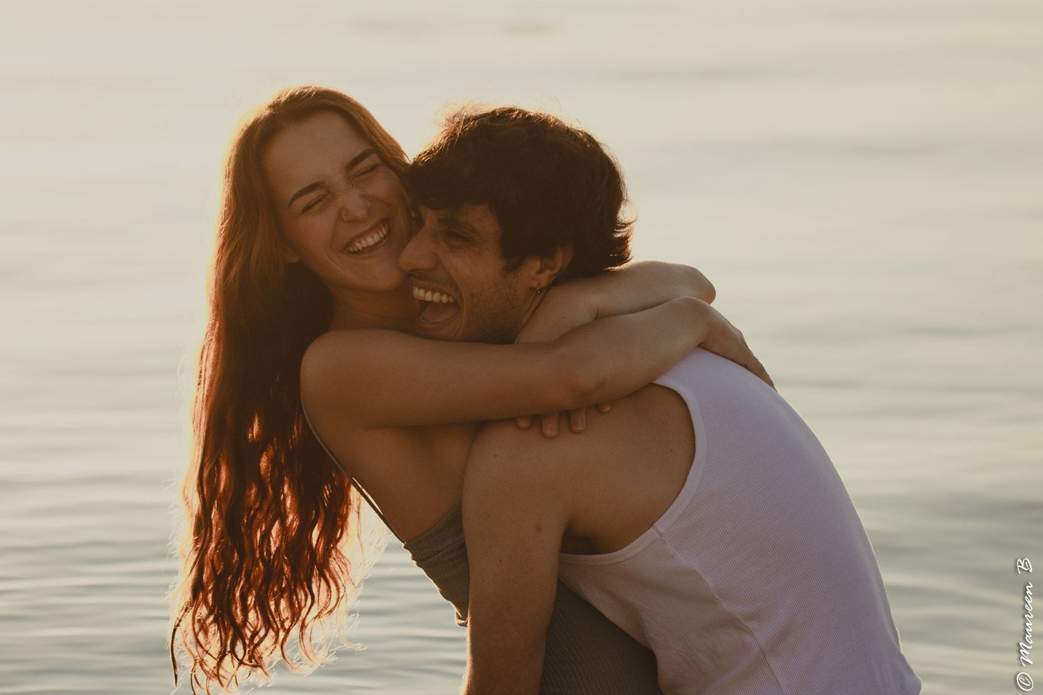 Joyful couple laughing and embracing on a sunlit beach at golden hour by the sea.