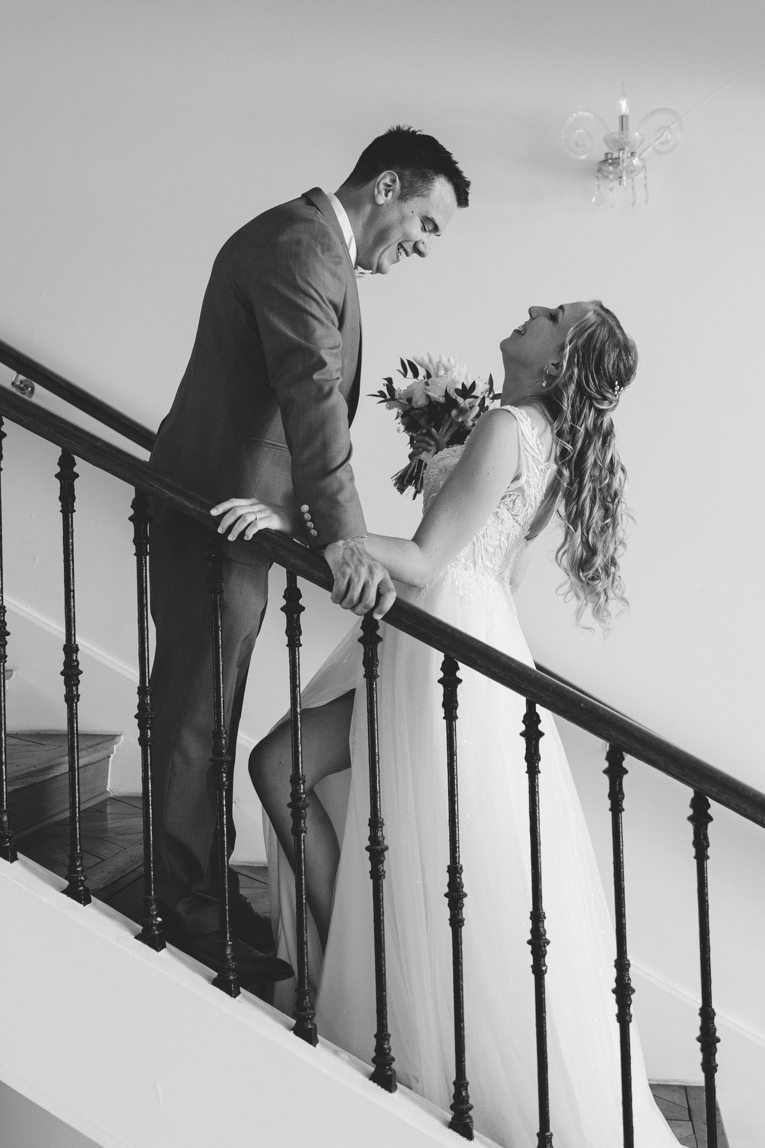 A black and white photo of a smiling bride and groom on a staircase with a bouquet.