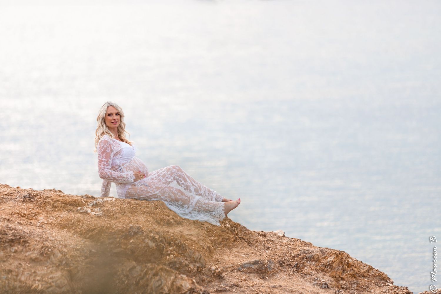 Pregnant woman in white lace dress posing gracefully on rocky coastal cliff by shimmering water.