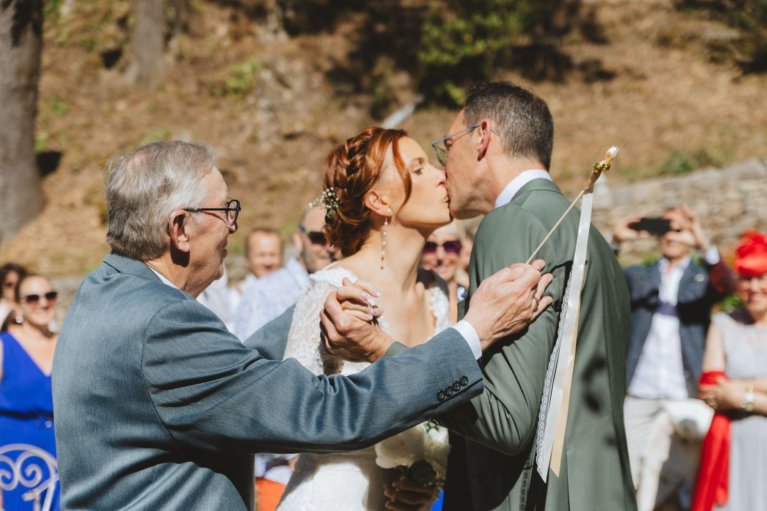 Bride and groom share a first kiss at outdoor wedding ceremony while elderly man watches joyfully.