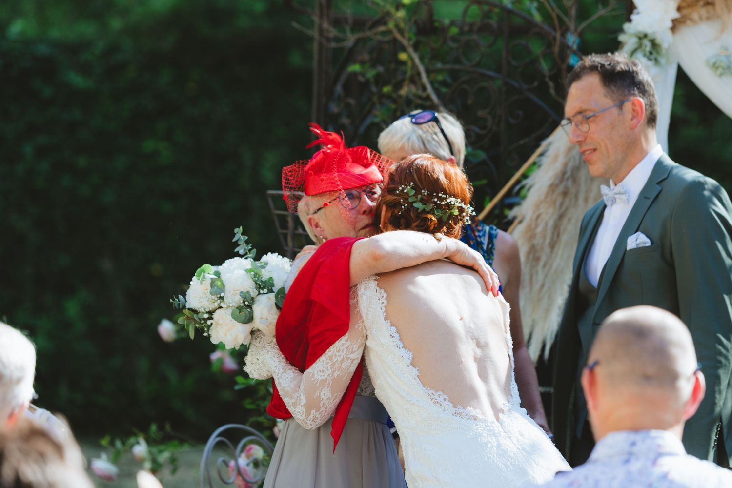 Bride in white gown hugging guest in red hat and cape at outdoor wedding ceremony with groom in green suit nearby.