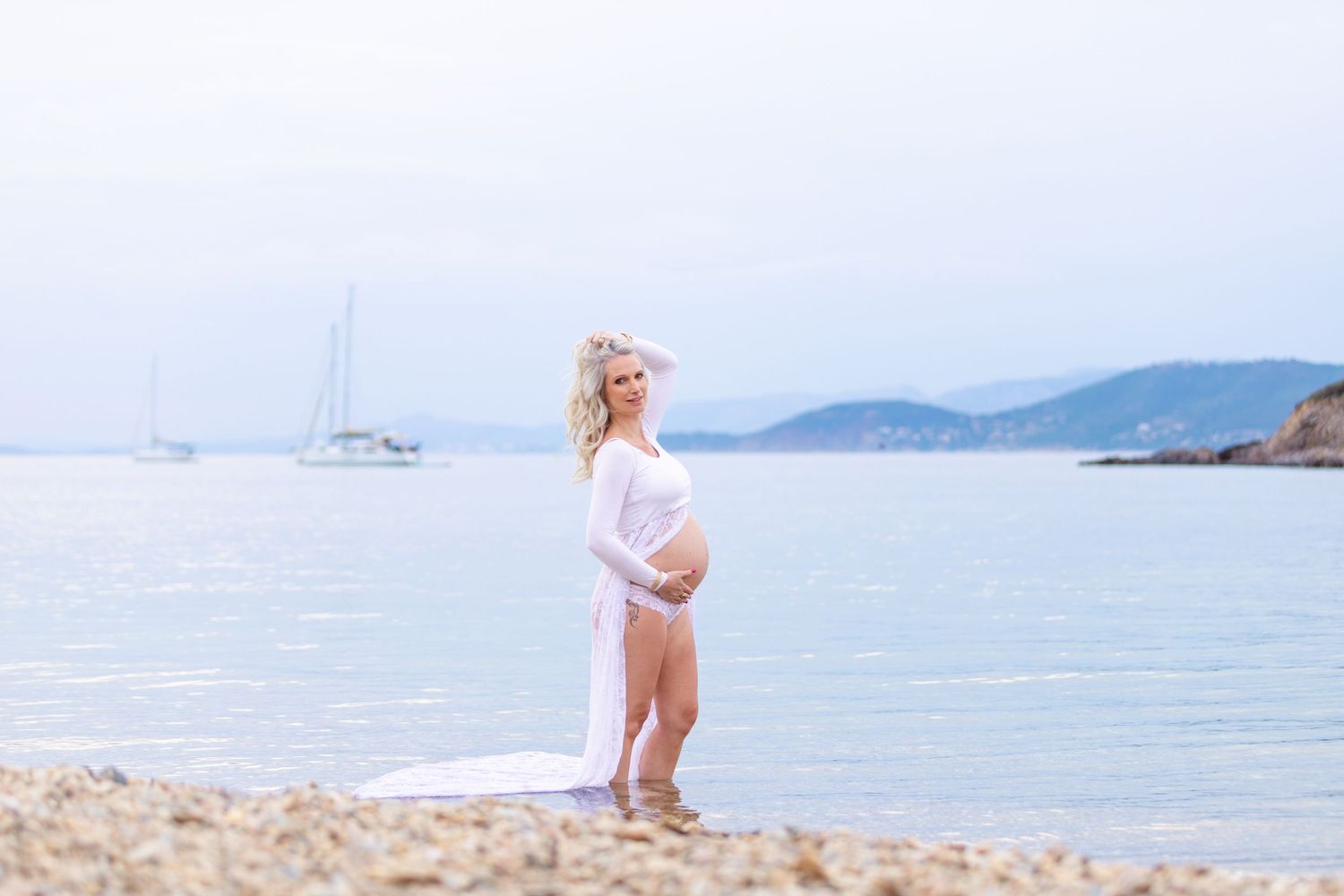 Pregnant woman in white sheer dress on beach, holding baby bump, sailboat and mountains in background.