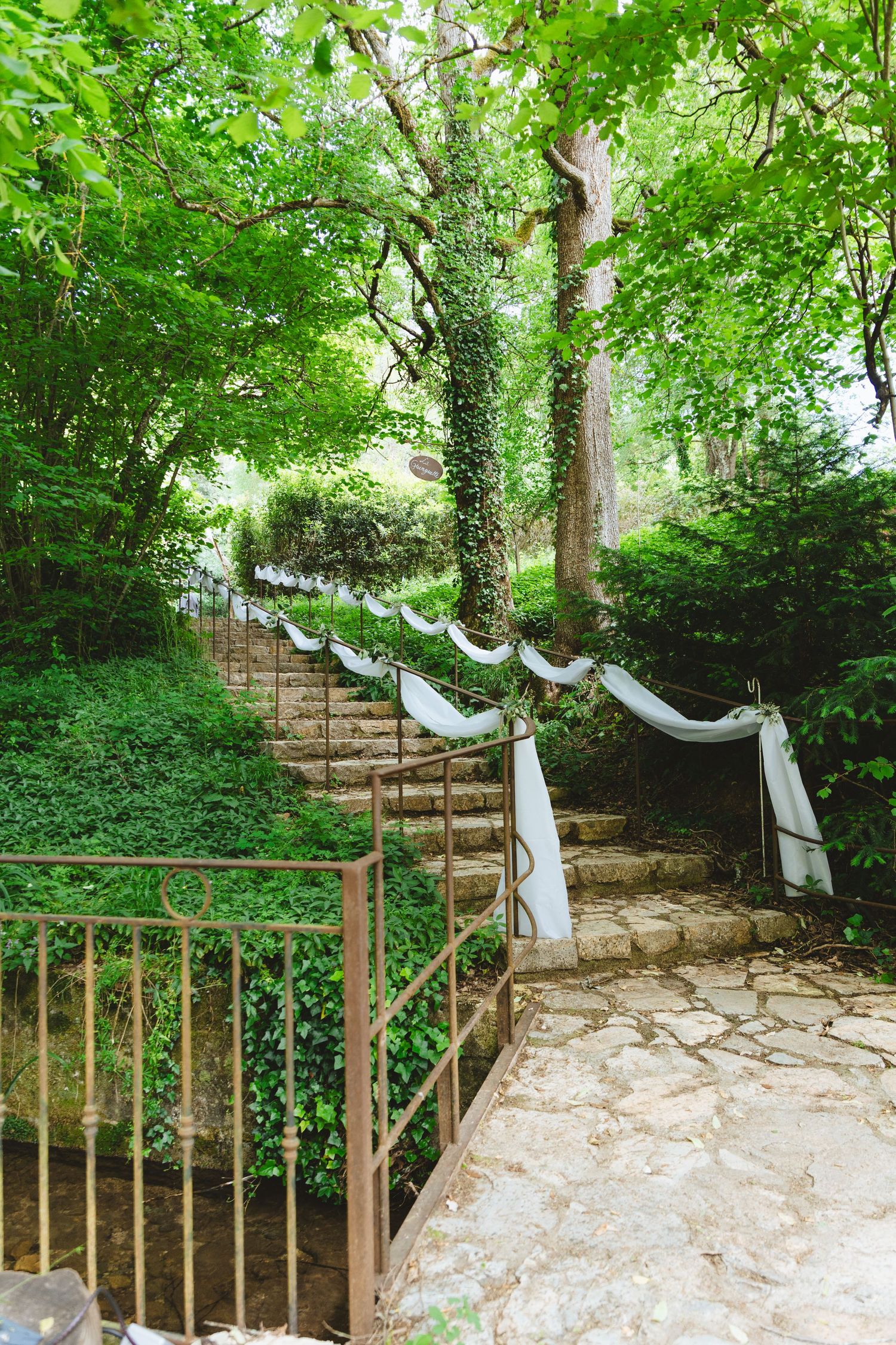 Stone staircase decorated with white fabric draped on railings, surrounded by lush green trees and ivy in a garden setting.