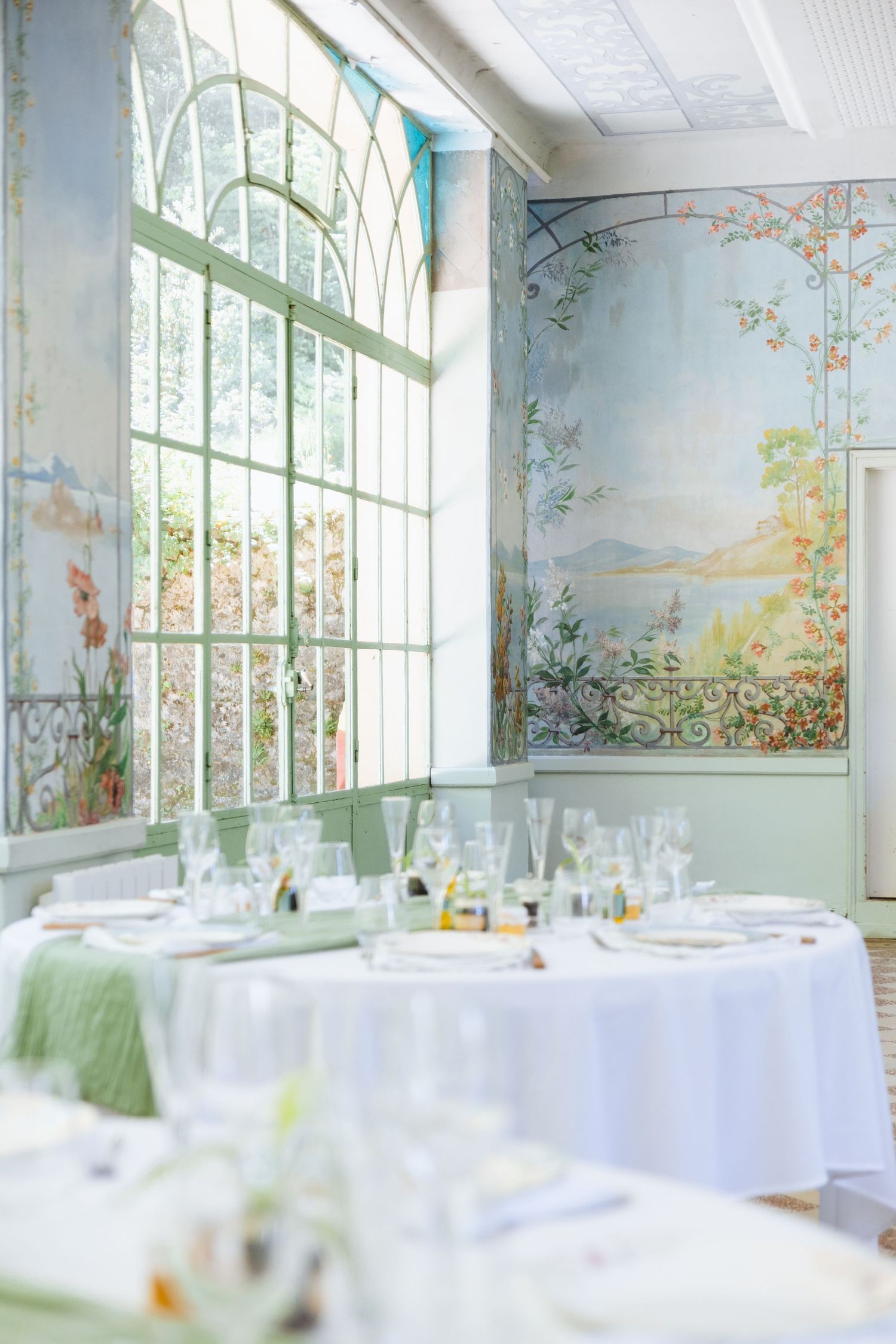 Elegant dining room with floral murals, arched windows, and white-draped tables set for a formal event.