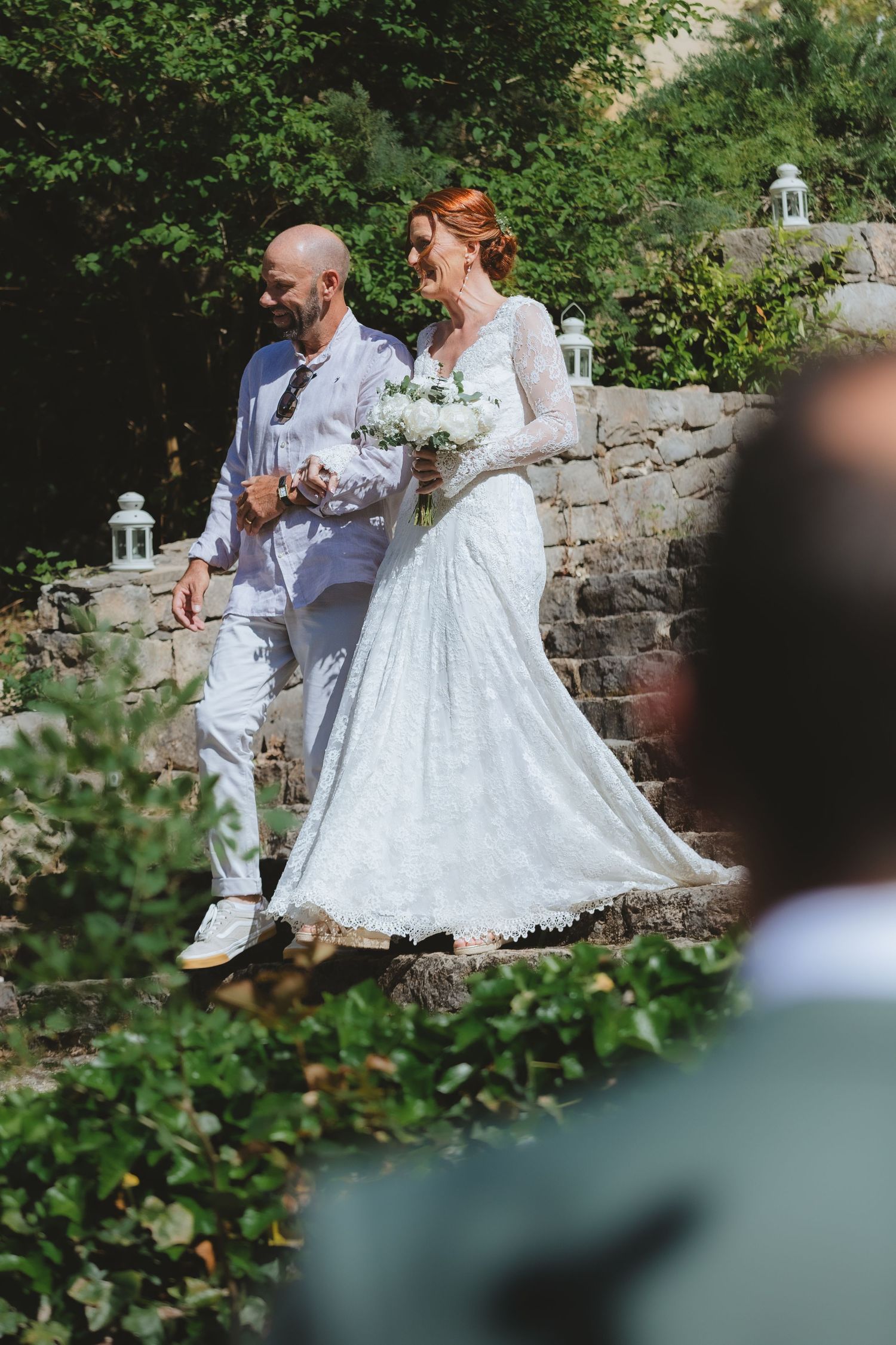 Bride in lace gown holding white bouquet walks with bald groom in white suit through lush garden wedding venue.