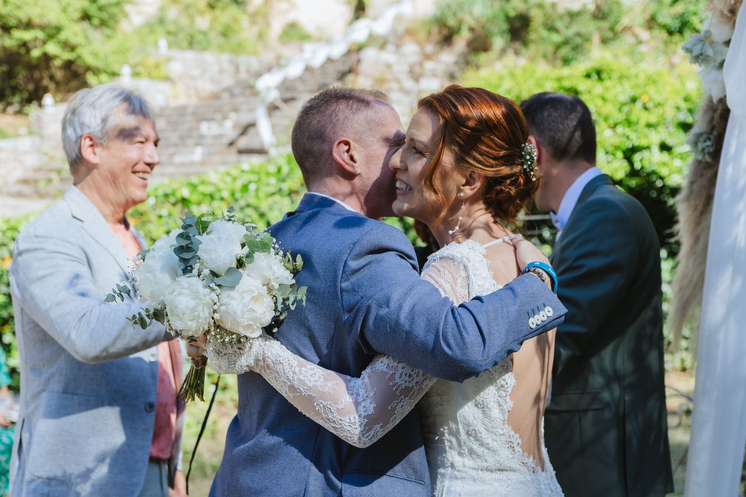 Bride and groom share first kiss at outdoor wedding ceremony while smiling officiant and guest look on.