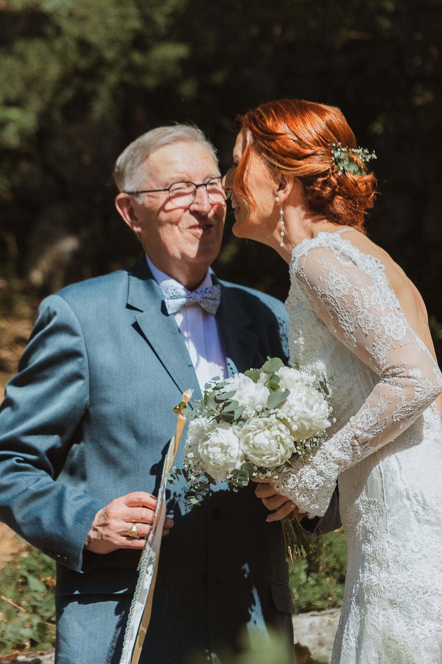 Bride in lace gown with white bouquet sharing a tender moment with elderly man in blue suit at outdoor wedding.