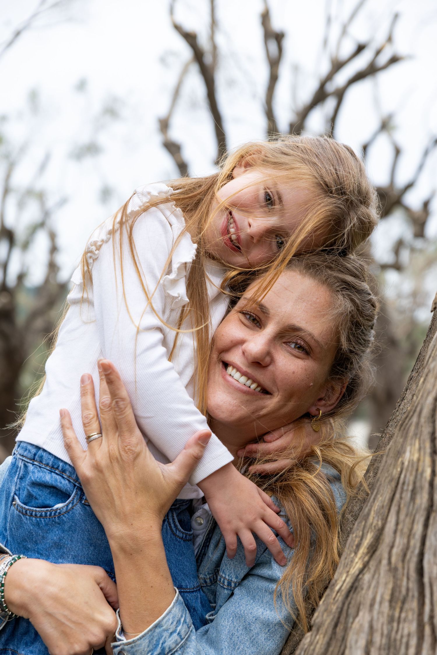 A smiling mother and daughter share a joyful embrace leaning against a tree outdoors.