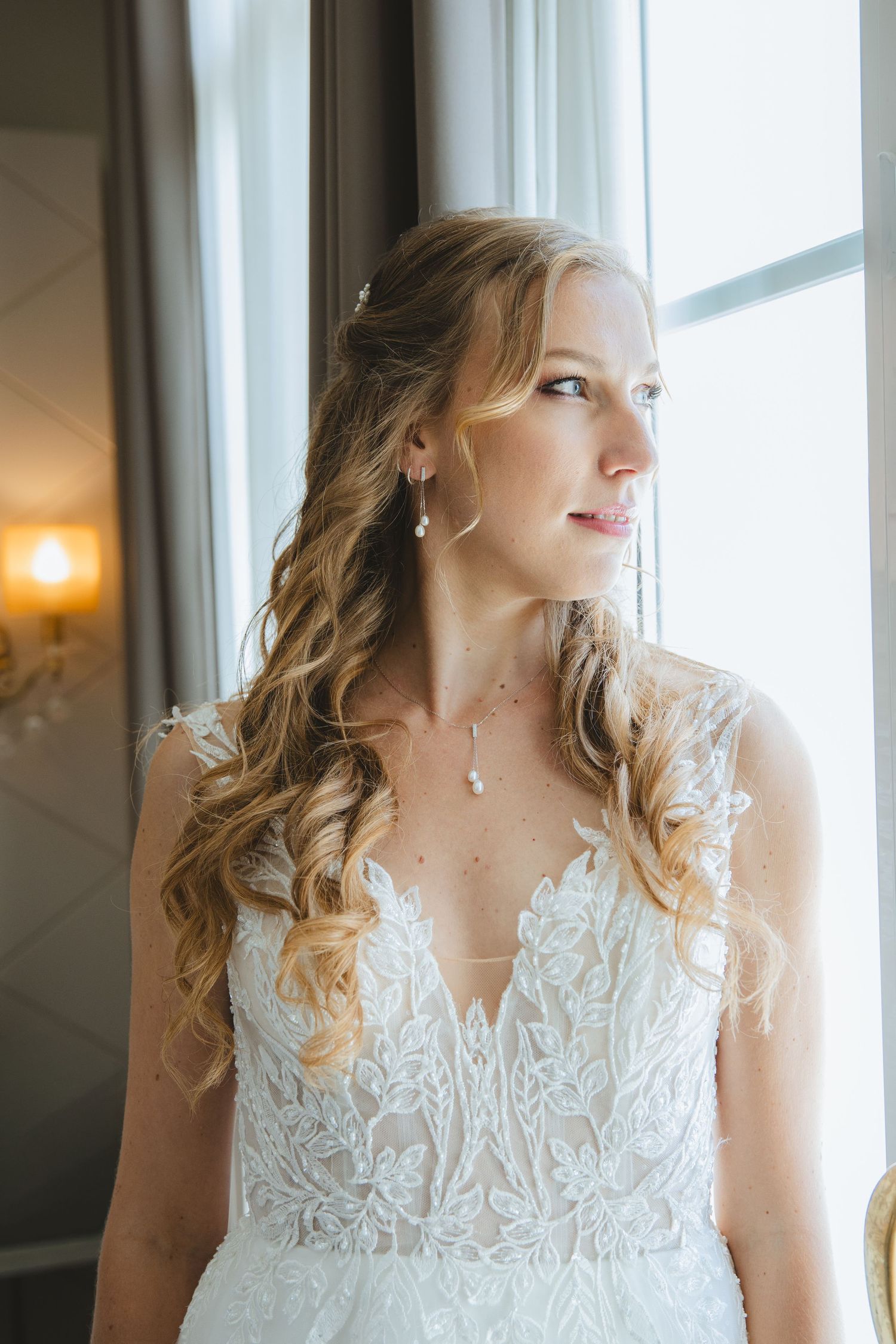 Elegant bride in lace V-neck gown with curled hair gazing out window in soft natural light.