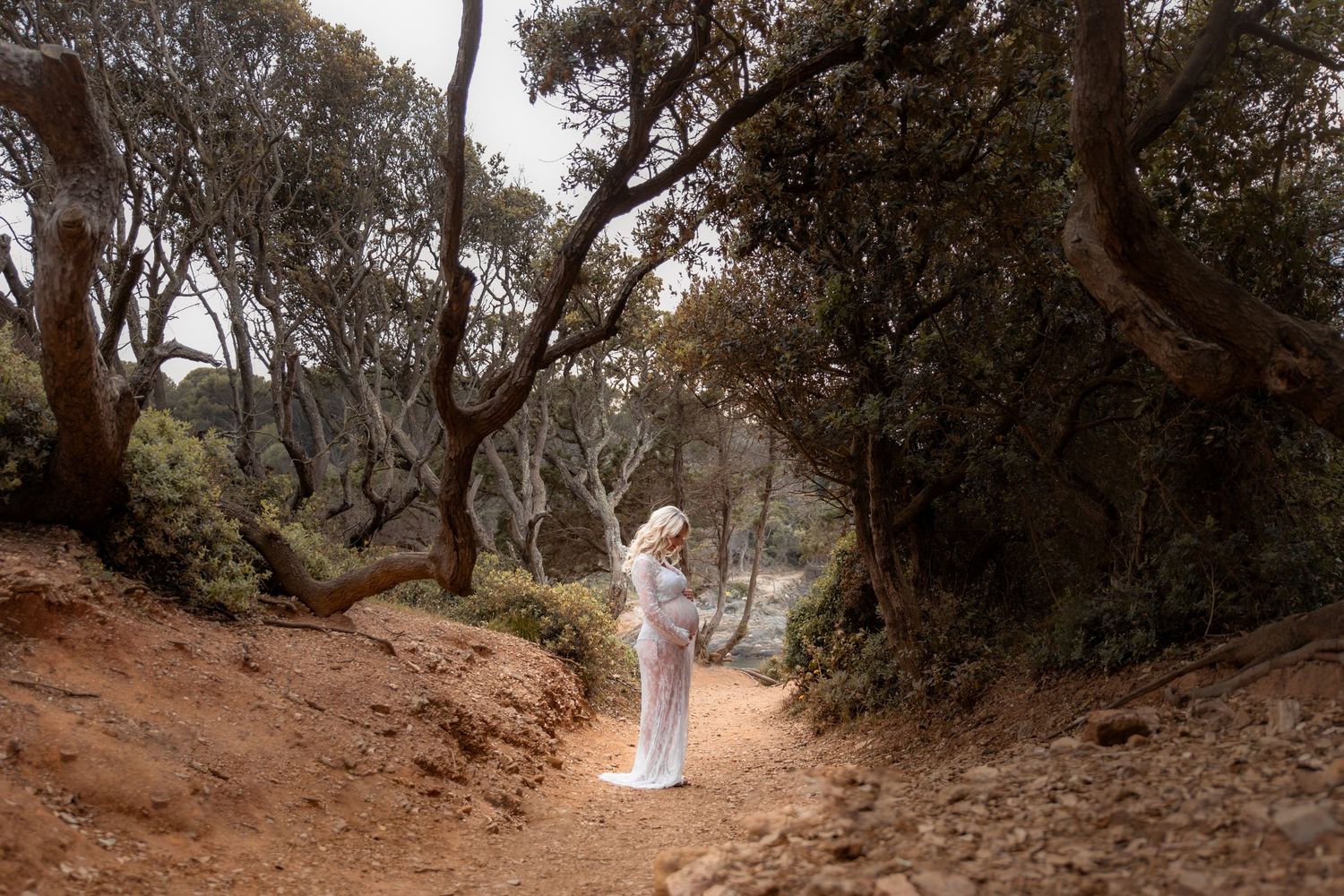 Pregnant woman in white gown poses on a forest trail surrounded by twisted trees and red earth.