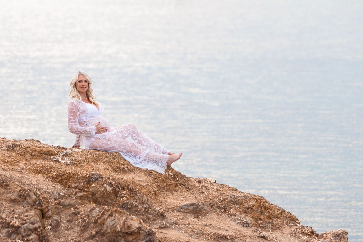 Pregnant woman in white lace dress sitting on rocky cliff overlooking sparkling ocean water.