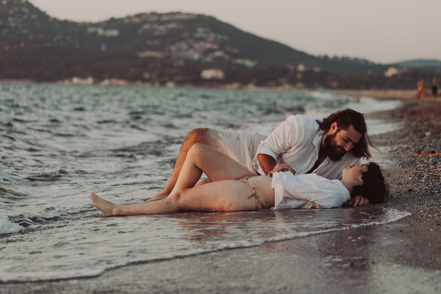 Romantic couple lying together on a sandy beach shore at sunset with mountains in the background.
