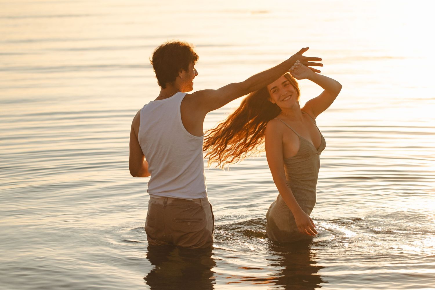 Couple dancing together in shallow ocean water during golden sunset, woman with long flowing hair, romantic beach scene.